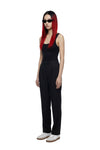 A model with long red hair and black sunglasses stands in profile wearing a black sleeveless top and black wide-leg trousers, paired with white sneakers, against a plain white background.