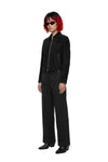 A model with short red hair and black sunglasses stands in a studio wearing a black zip-front jacket, black wide-leg trousers, and black shoes against a plain white background.