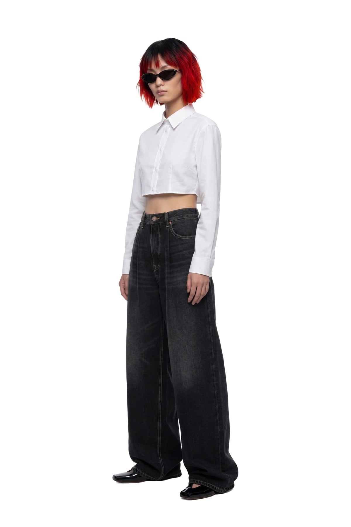A model with short red hair and black sunglasses stands in profile wearing a white cropped long-sleeve button-up shirt and very wide-leg black jeans against a plain white background.