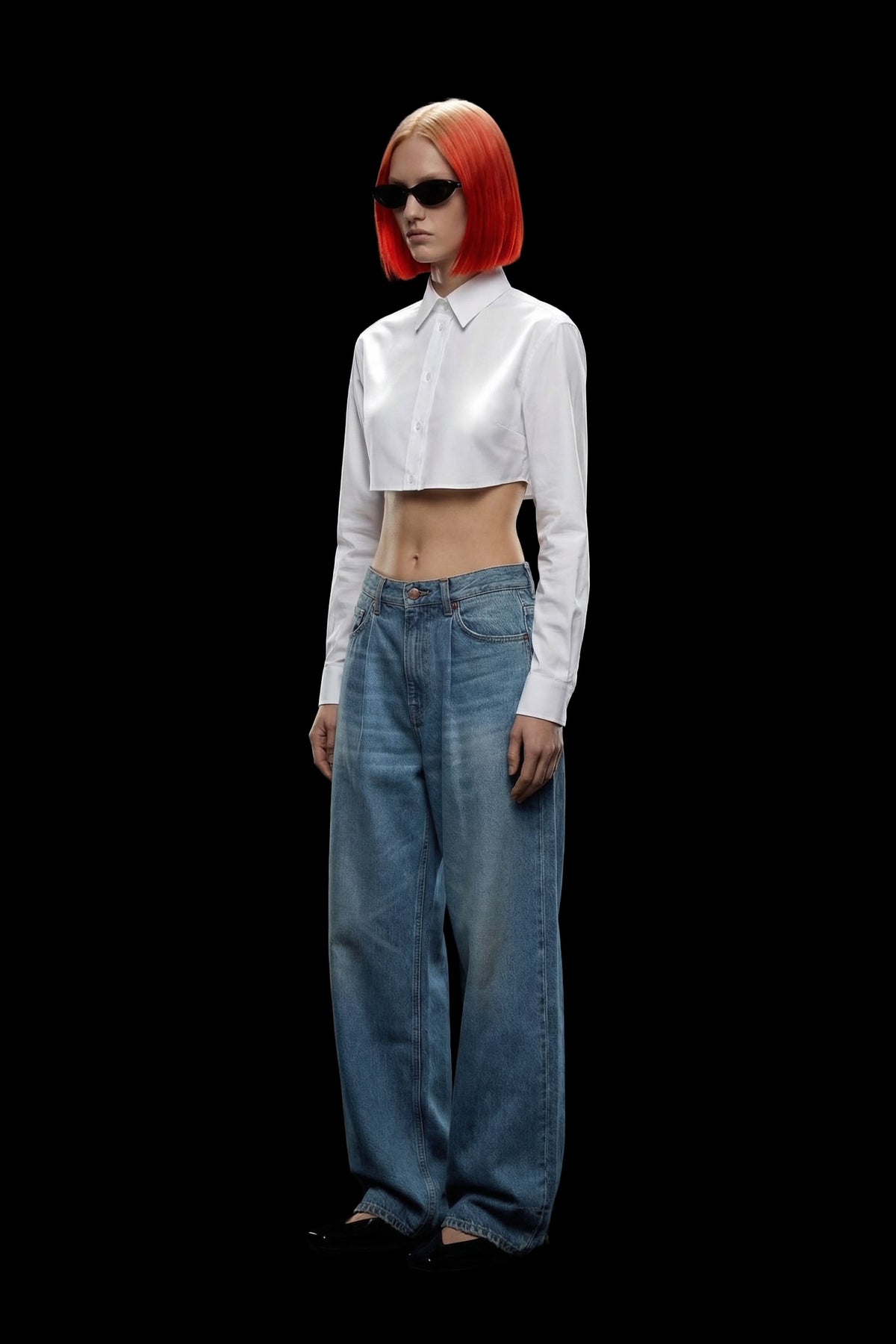 A model with bright orange hair and black sunglasses stands against a solid black background, wearing a white long-sleeve cropped button-up shirt and loose blue jeans.