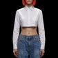 A model with bright orange hair and black sunglasses wears a white long-sleeve cropped button-up shirt paired with high-waisted blue jeans, set against a solid black background.