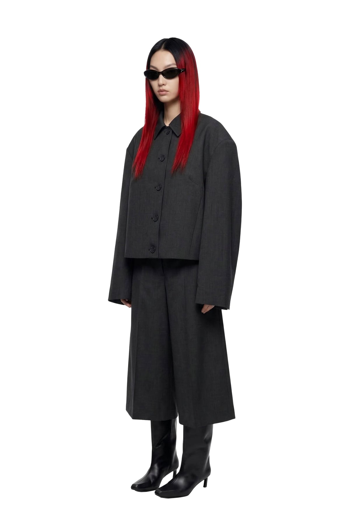 A model with long red hair and black sunglasses stands against a white background wearing a dark gray cropped button-up jacket, matching wide-leg trousers, black knee-high boots, and black leather loafers.