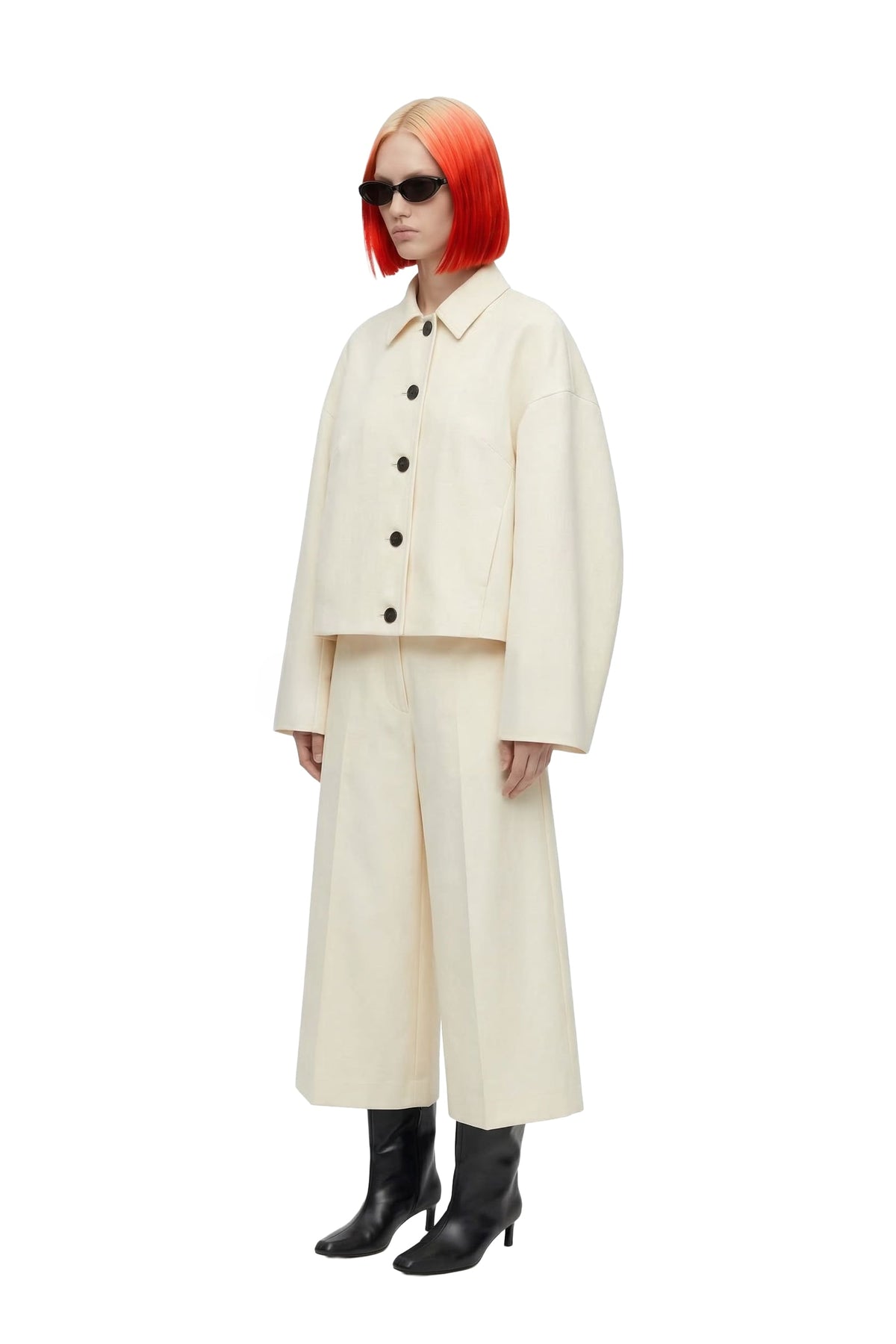 A model with vibrant orange hair and black sunglasses wearing a cream-colored cropped jacket and matching wide-leg trousers, paired with black leather boots, against a solid white background.