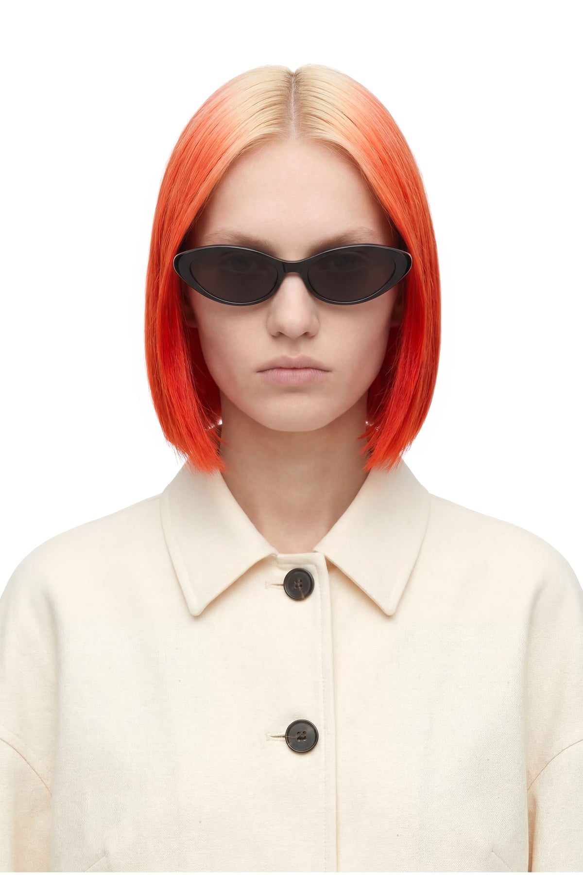 A model with a vibrant orange-to-blonde ombre bob haircut wearing black cat-eye sunglasses and a cream-colored collared jacket, set against a plain white background.
