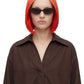 A model with a short, straight, bright orange-red ombre blonde wig and black sunglasses, wearing an oversized dark brown button-up shirt against a plain white background.