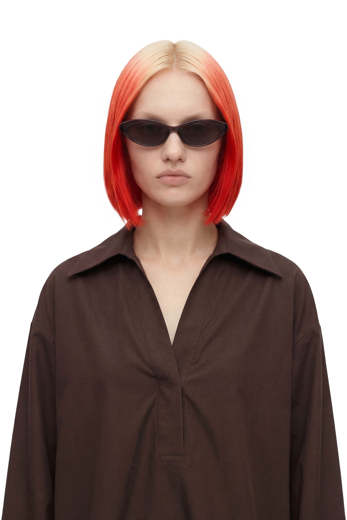 A model with a short, straight, bright orange-red ombre blonde wig and black sunglasses, wearing an oversized dark brown button-up shirt against a plain white background.