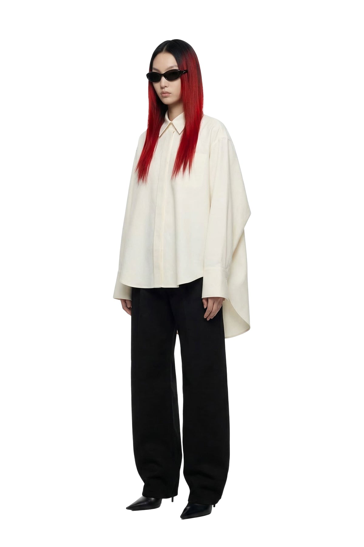 A model with long red hair and black sunglasses stands against a white background, wearing a loose off-white oversized button-down shirt, black wide-leg trousers, and black pointed shoes.