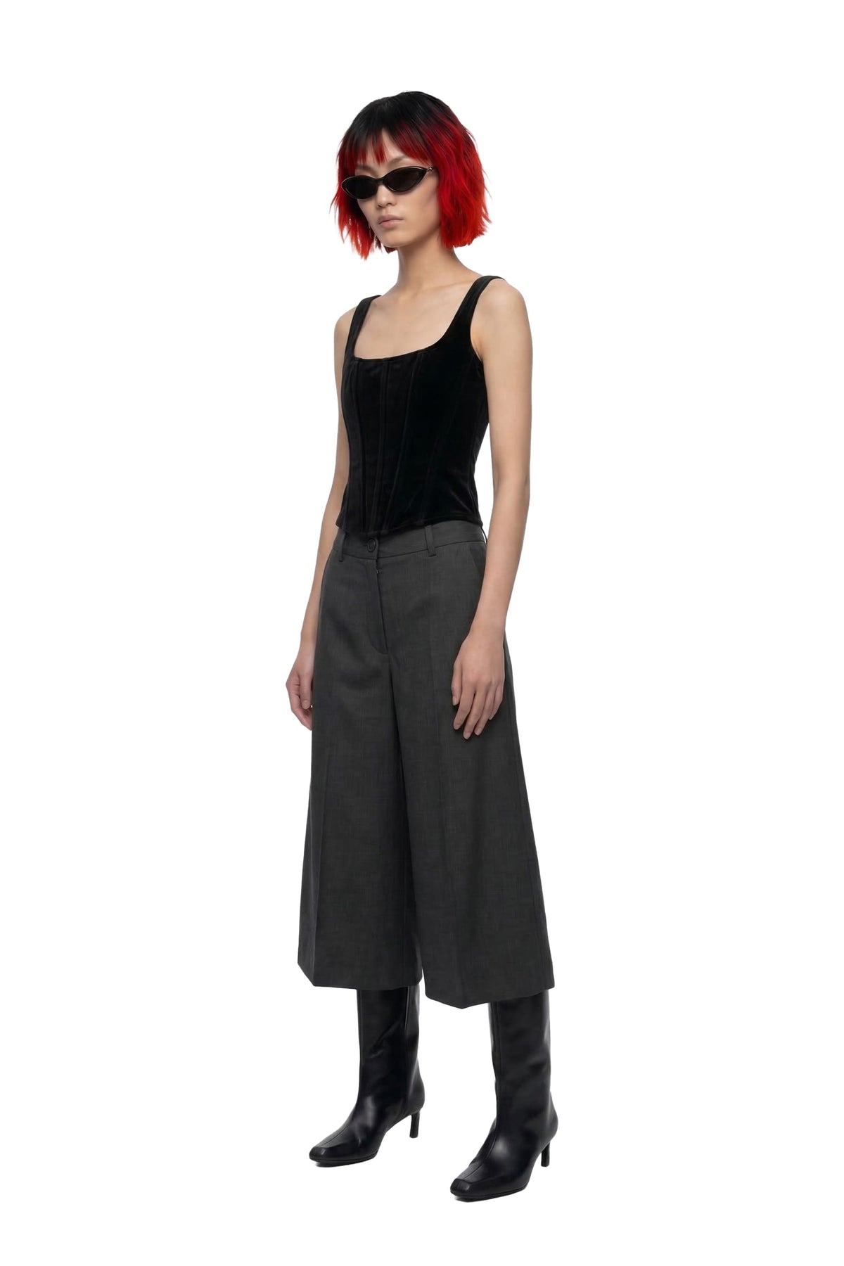 A model with a red and black bob haircut wearing a black velvet tank top, dark gray wide-leg cropped trousers, and black knee-high boots, set against a plain white background.
