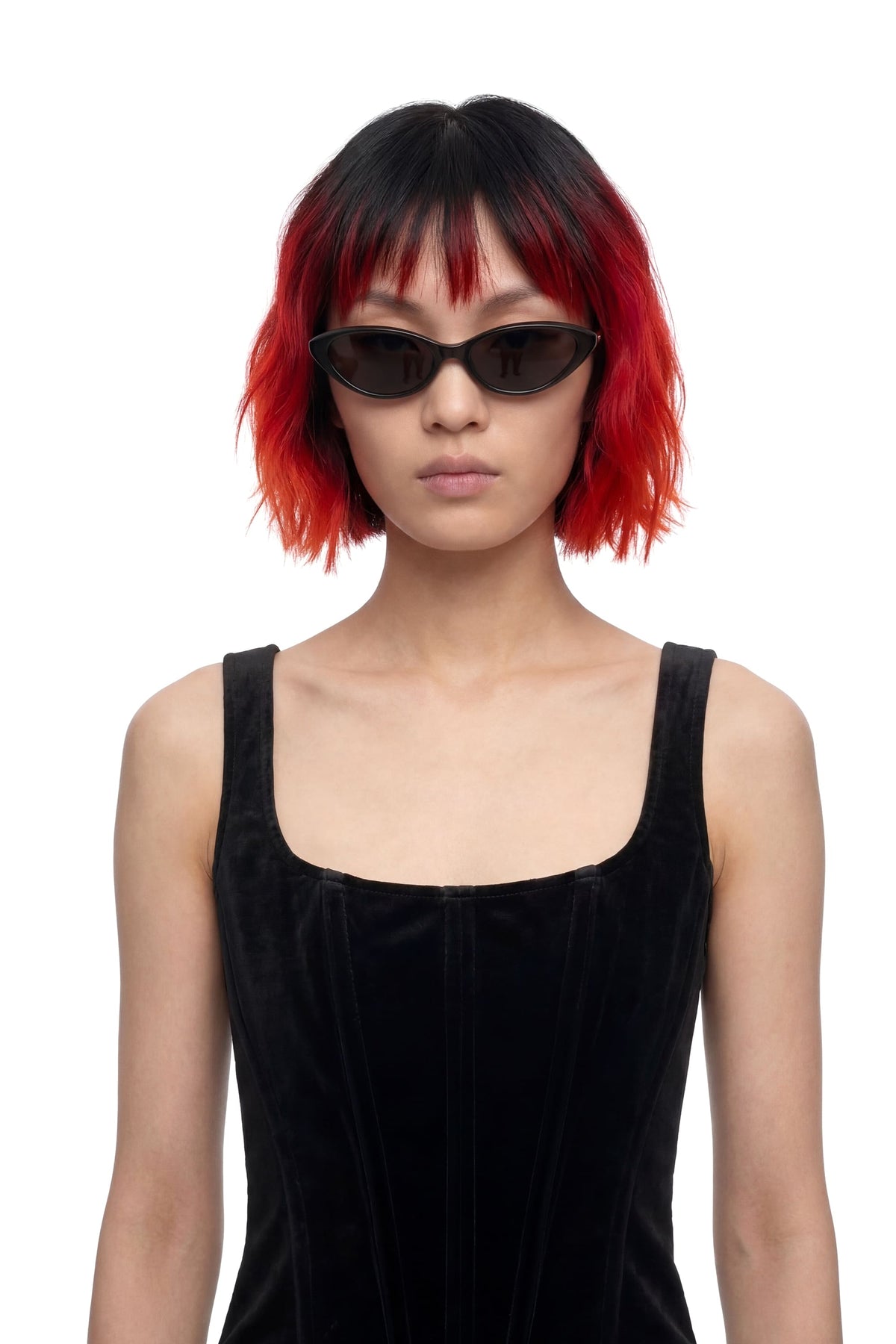 A model with a short black and vibrant red ombre wig, wearing black cat-eye sunglasses and a black velvet sleeveless top against a plain white background.
