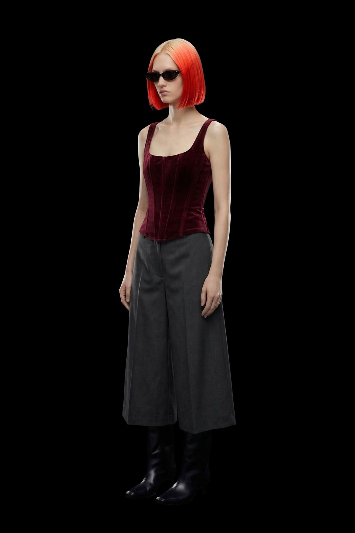 A digital render of a female model with vibrant orange hair and sunglasses, wearing a burgundy velvet corset top, dark gray wide-leg cropped pants, and black boots against a solid black background.