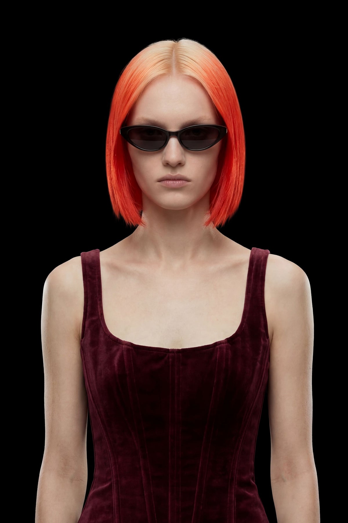 A model with a vibrant orange-to-blonde ombre bob haircut, wearing black cat-eye sunglasses and a deep burgundy velvet corset top, set against a solid black background.