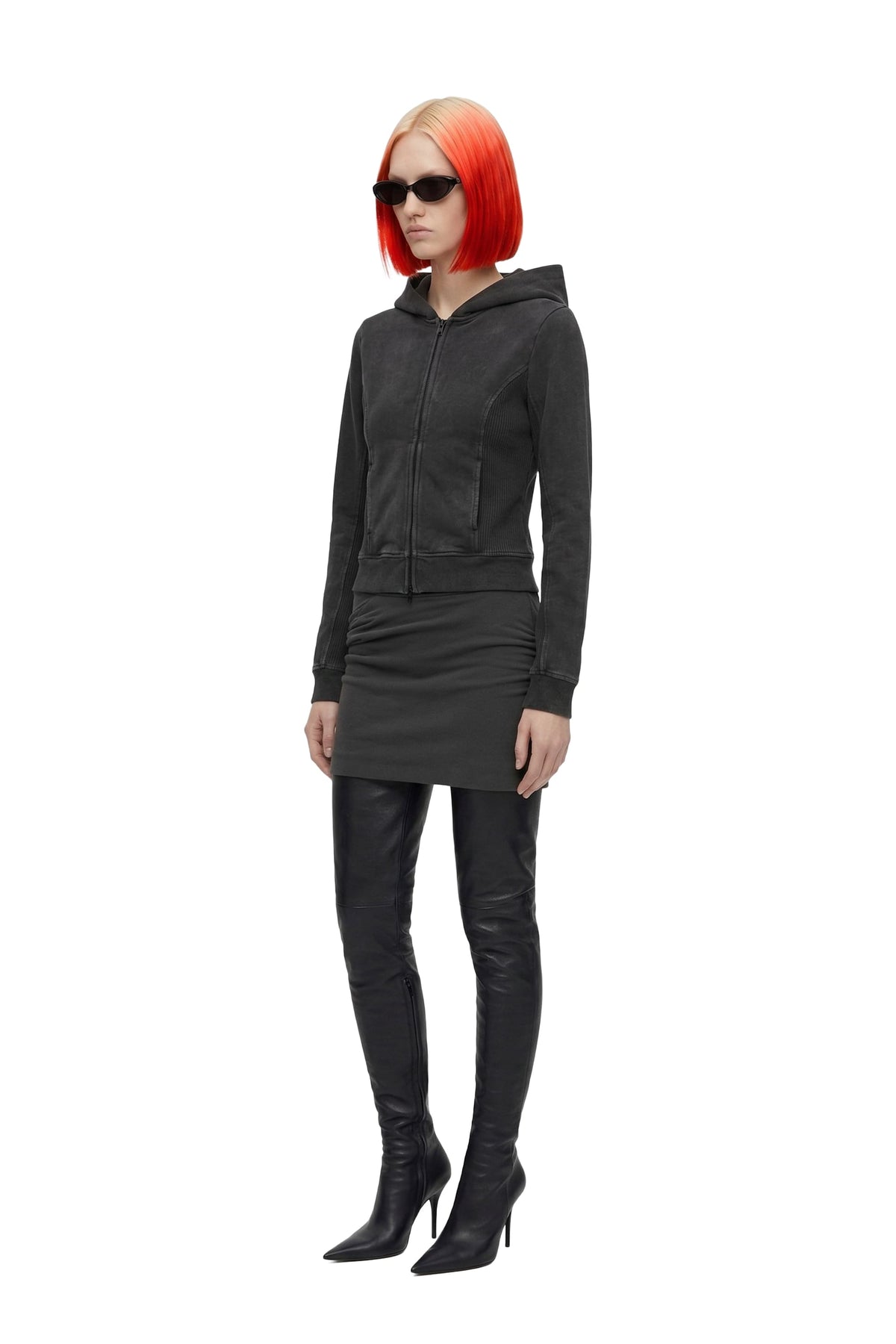 A model with a bright orange-red bob haircut and black sunglasses is shown in profile wearing a black zip-up hooded sweatshirt, a matching black mini skirt, black faux-leather pants, and black high-heeled boots against a plain white background.