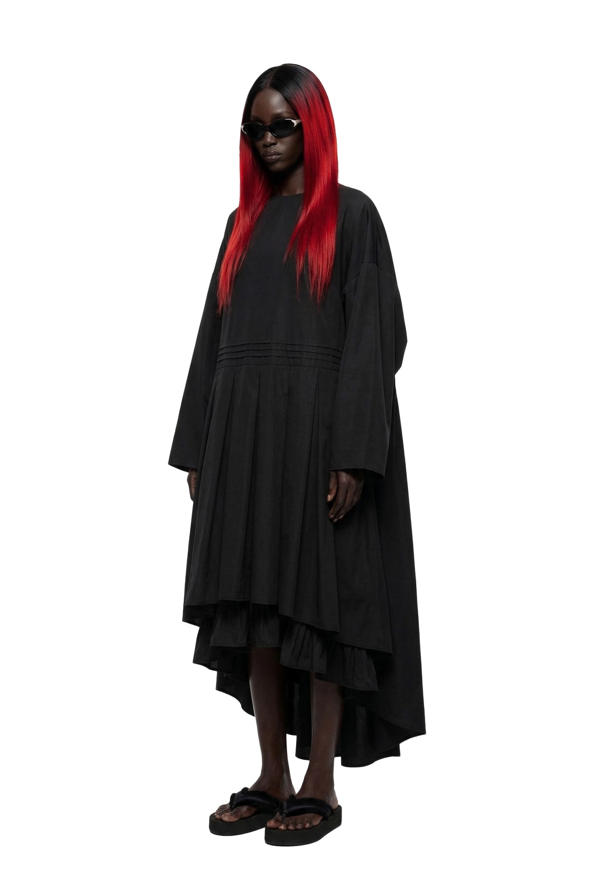 A model with long black hair and bright red streaks wearing a loose, oversized black long-sleeve dress with a tiered ruffled hem, black sunglasses, and black sandals, set against a plain white background.
