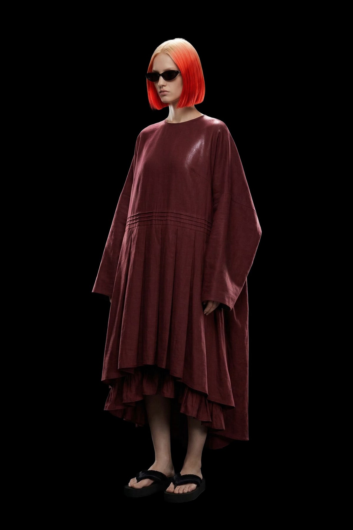 A model with a bright orange-red bob haircut and sunglasses wearing a dark reddish-brown high-low dress with wide sleeves, standing against a solid black background.
