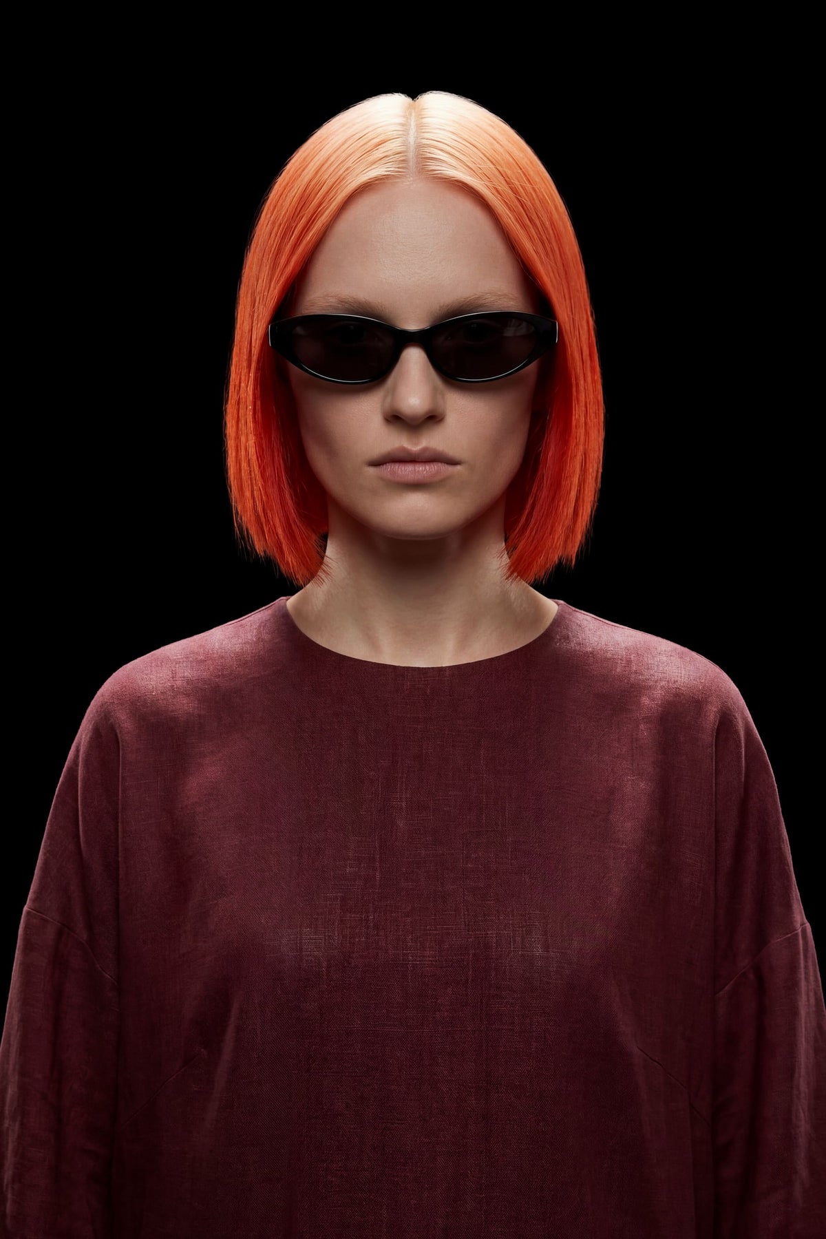 A model with vibrant orange hair wearing black cat-eye sunglasses and a textured burgundy top, set against a solid black background.