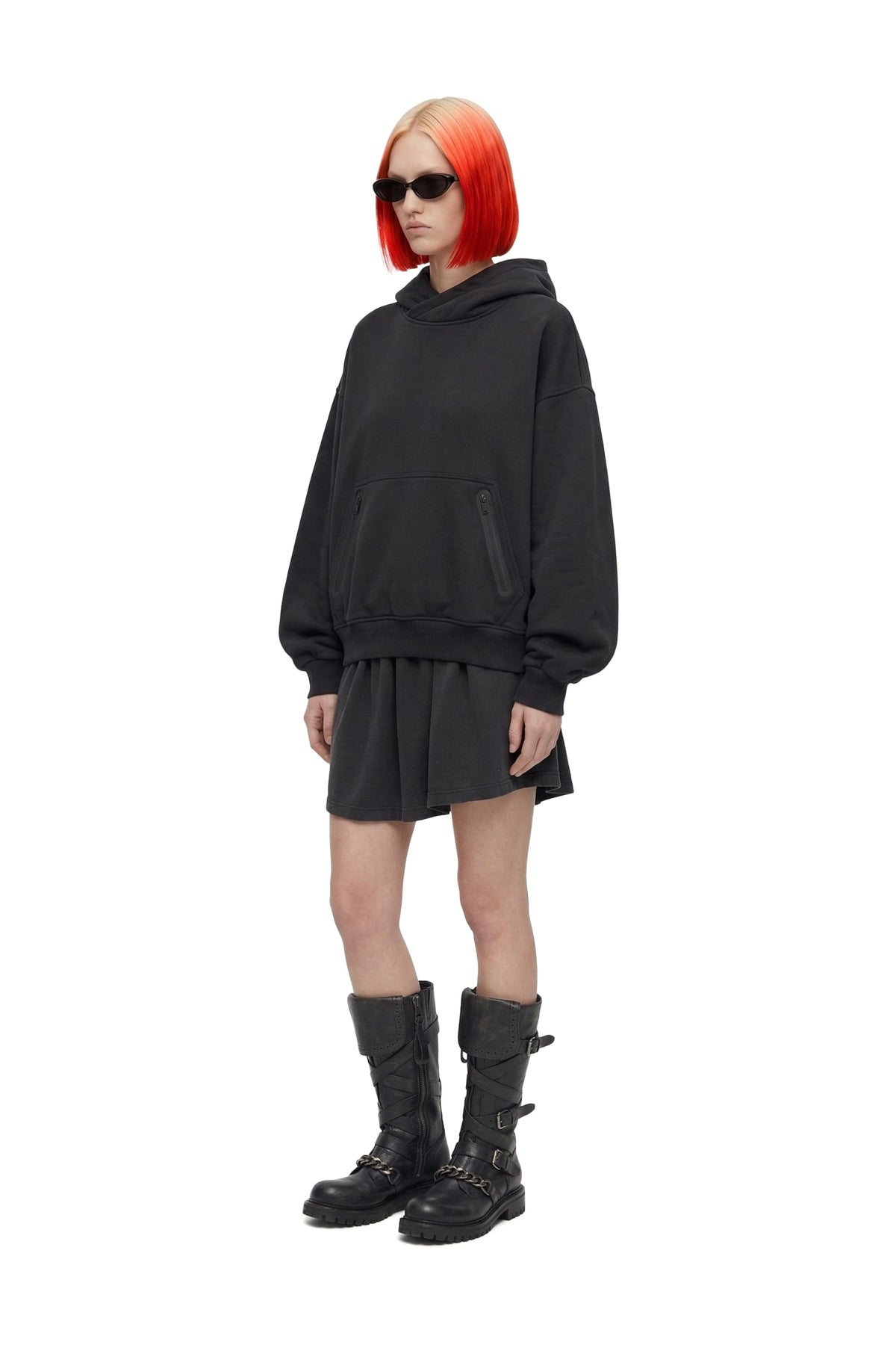 A model with bright orange hair and sunglasses wearing a black oversized hoodie dress, black mini skirt, and black combat boots against a white background.