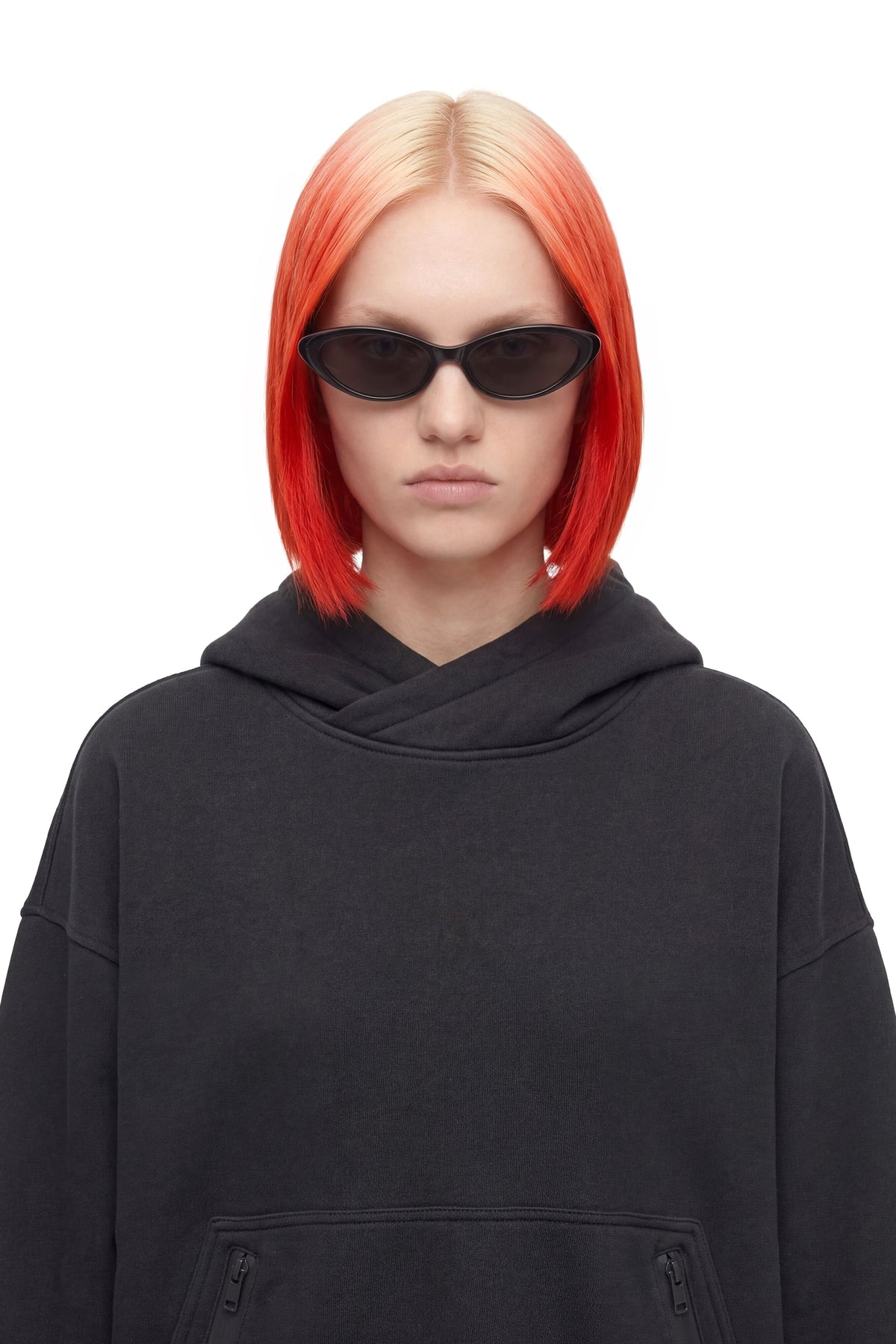 A model with vibrant red and blonde ombre hair wearing black cat-eye sunglasses and a black oversized hoodie, photographed against a solid white background.