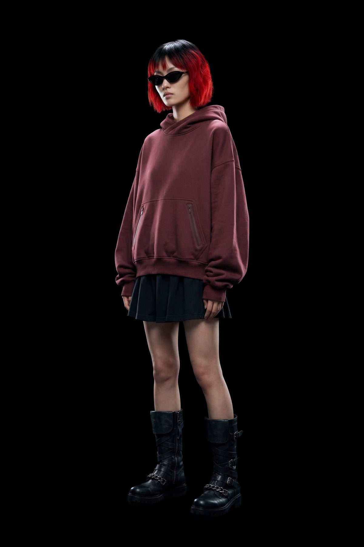 A model with a red and black bob haircut wearing a dark burgundy oversized hoodie, a black pleated mini skirt, black combat boots, and black sunglasses, standing against a solid black background.