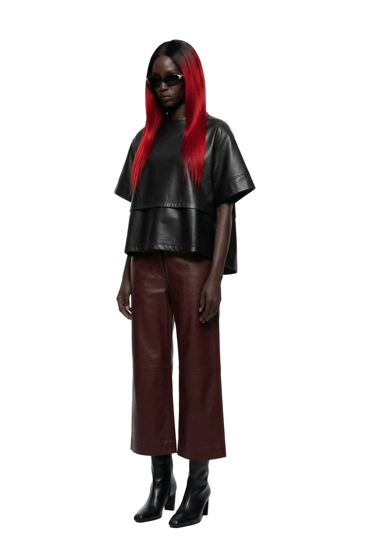 A model with long red hair wearing a black leather short-sleeve top and dark brown wide-leg leather pants, paired with black boots and sunglasses, against a plain white background.