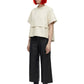 A model with bright orange hair and black sunglasses stands against a white background, wearing a cream-colored layered short-sleeve top, black wide-leg cropped pants, and black ankle boots.