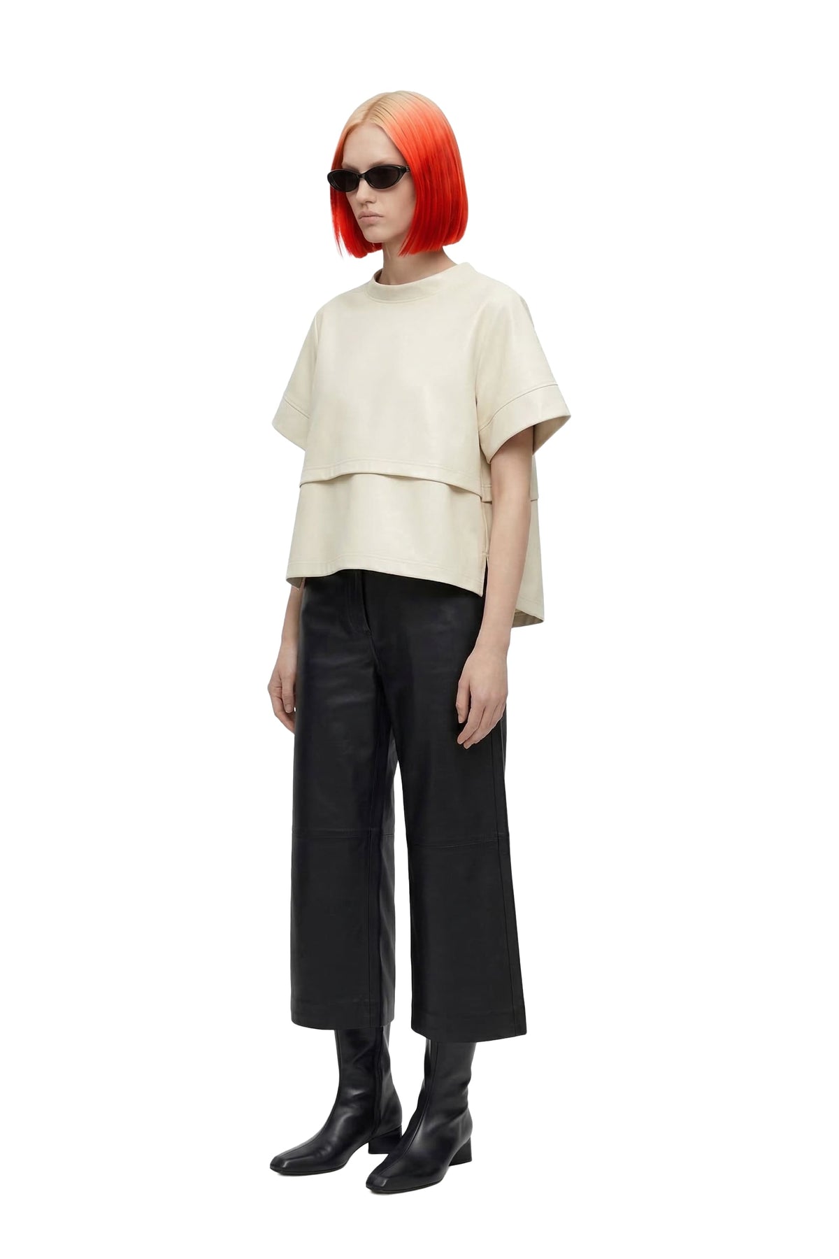 A model with bright orange hair and black sunglasses stands against a white background, wearing a cream-colored layered short-sleeve top, black wide-leg cropped pants, and black ankle boots.