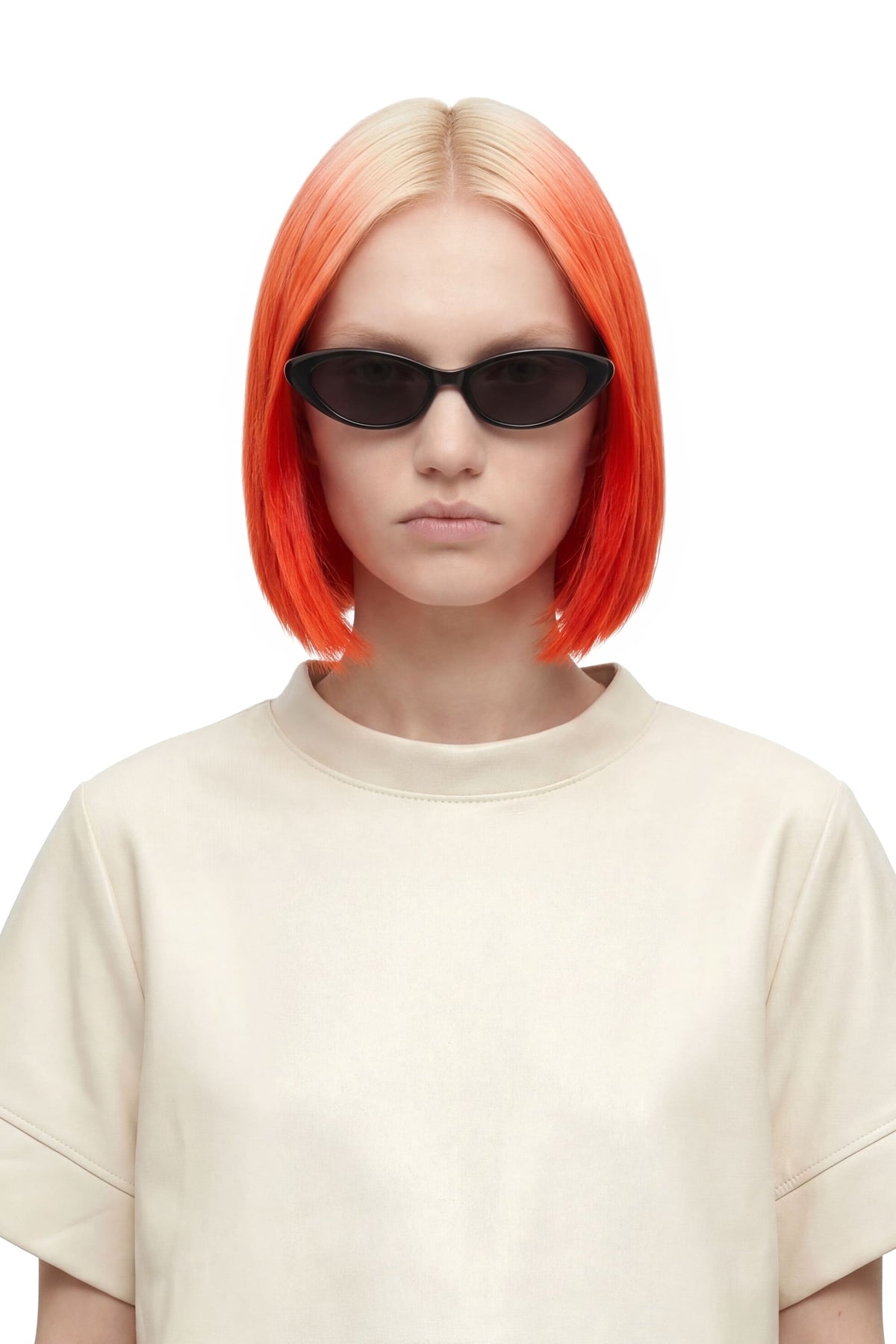 A model with a vibrant orange-to-blonde ombre bob haircut wearing black cat-eye sunglasses and a cream-colored oversized t-shirt, set against a plain white background.