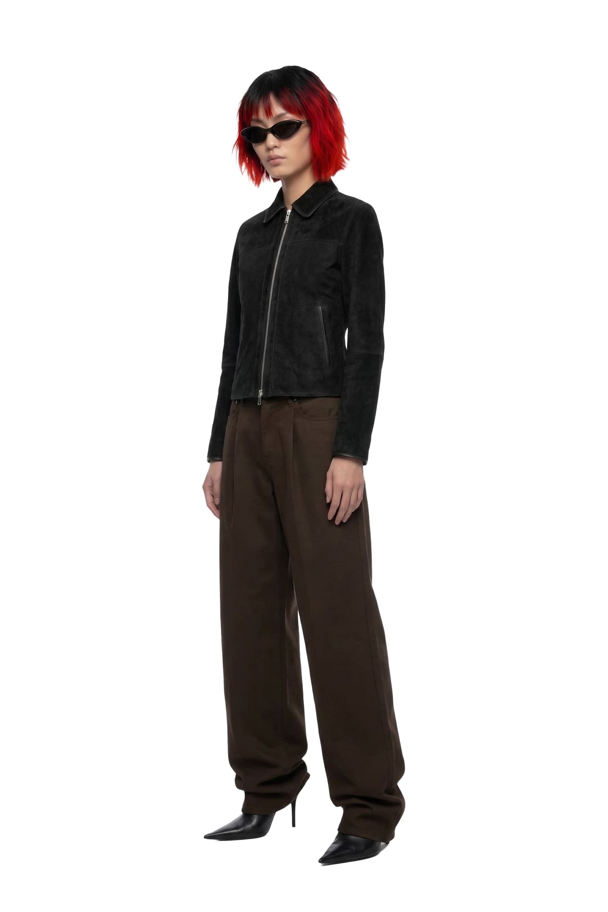 A model with short red hair and black sunglasses stands against a white background wearing a black cropped zip-up jacket, brown wide-leg pants, and black pointed-toe heels.