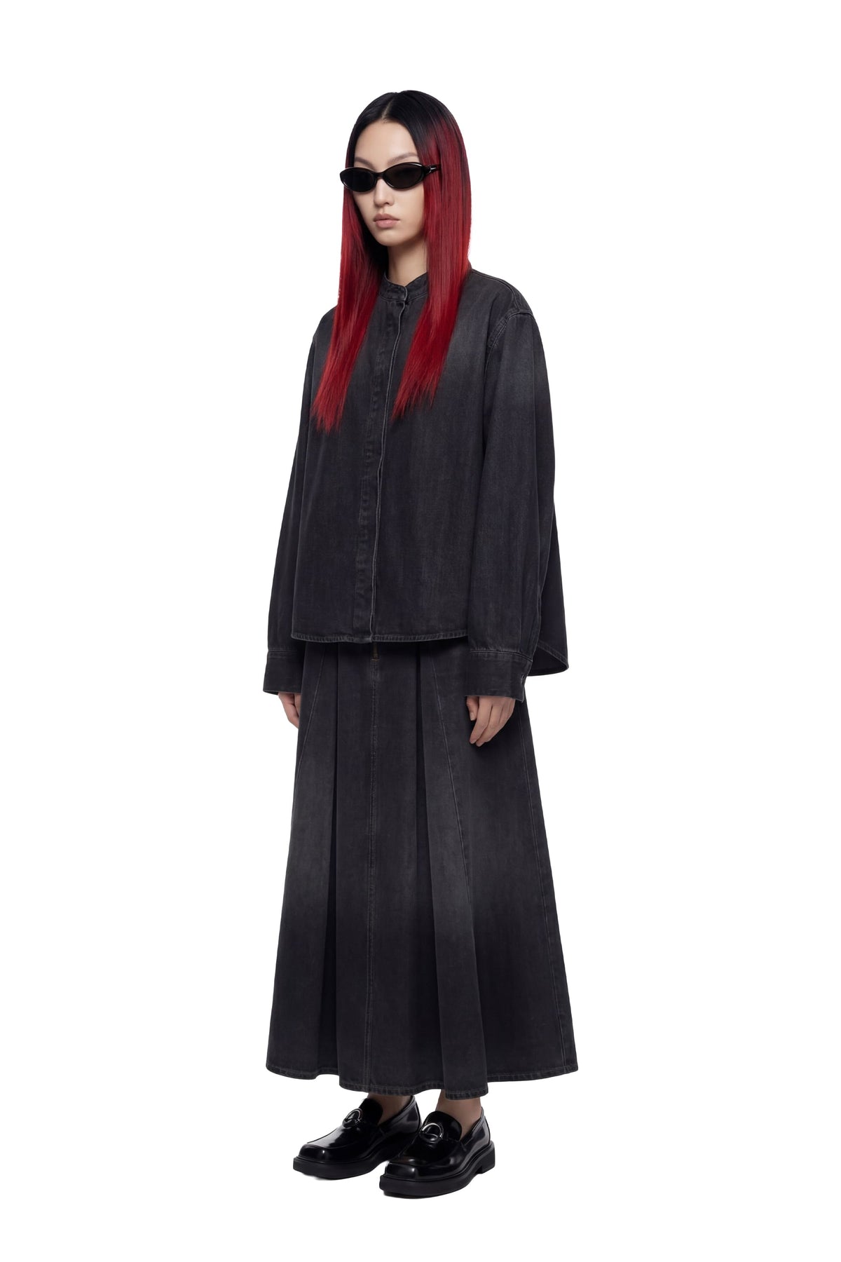A model with long red hair and black sunglasses stands against a white background wearing a dark charcoal gray two-piece outfit consisting of a cropped button-up jacket and a matching pleated maxi skirt, paired with black loafers.
