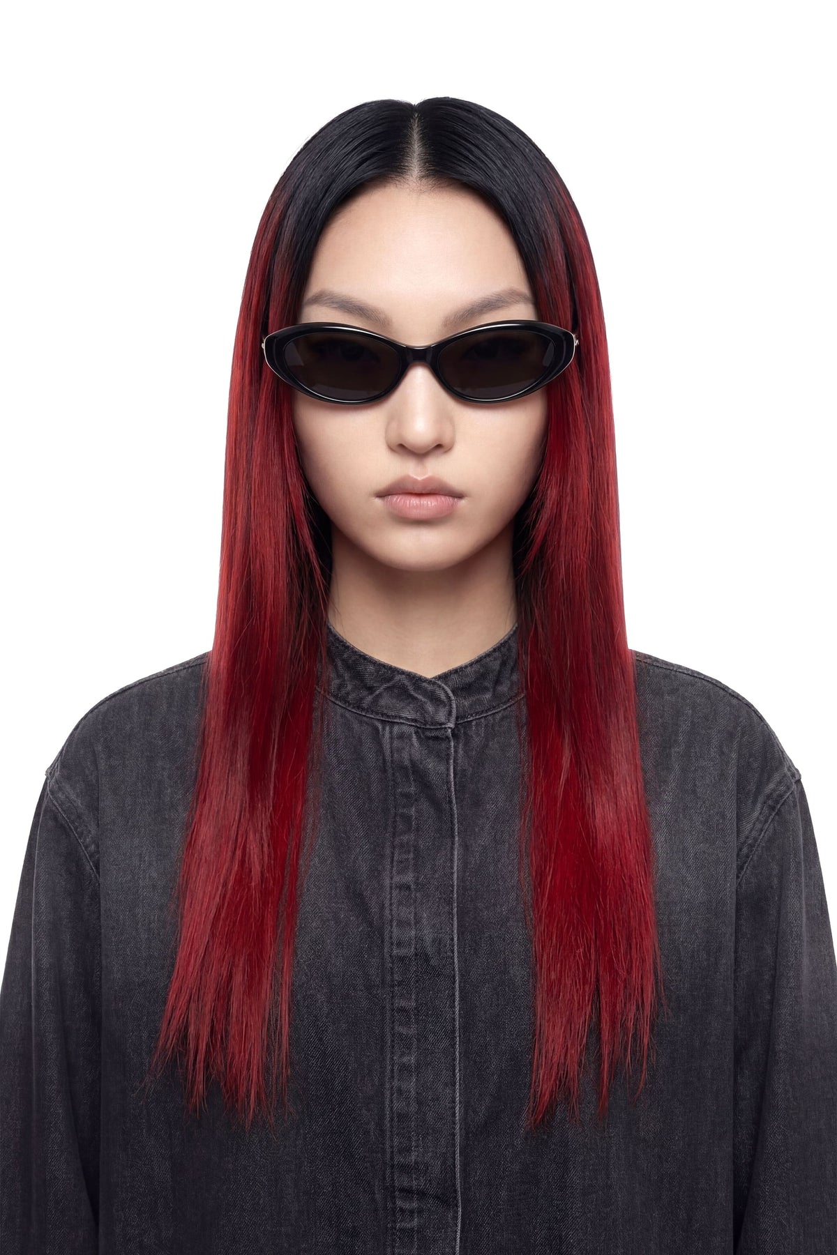 A model with long, straight black hair featuring vibrant red ombre ends, wearing black cat-eye sunglasses and a dark gray denim shirt against a solid white background.