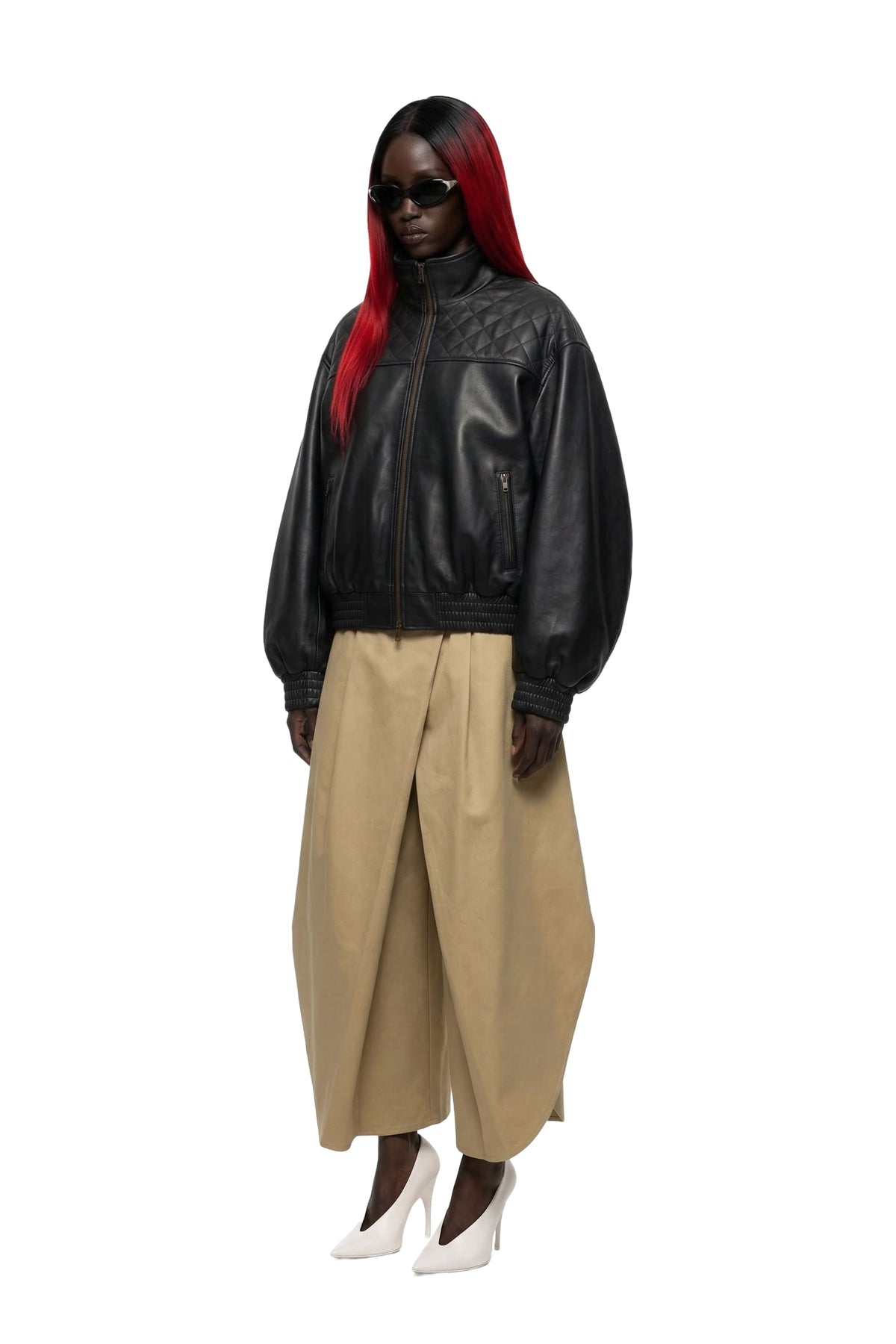 A model with long red hair wearing a black leather jacket, beige wide-leg pleated trousers, and white pointed-toe heels, photographed against a solid white background.