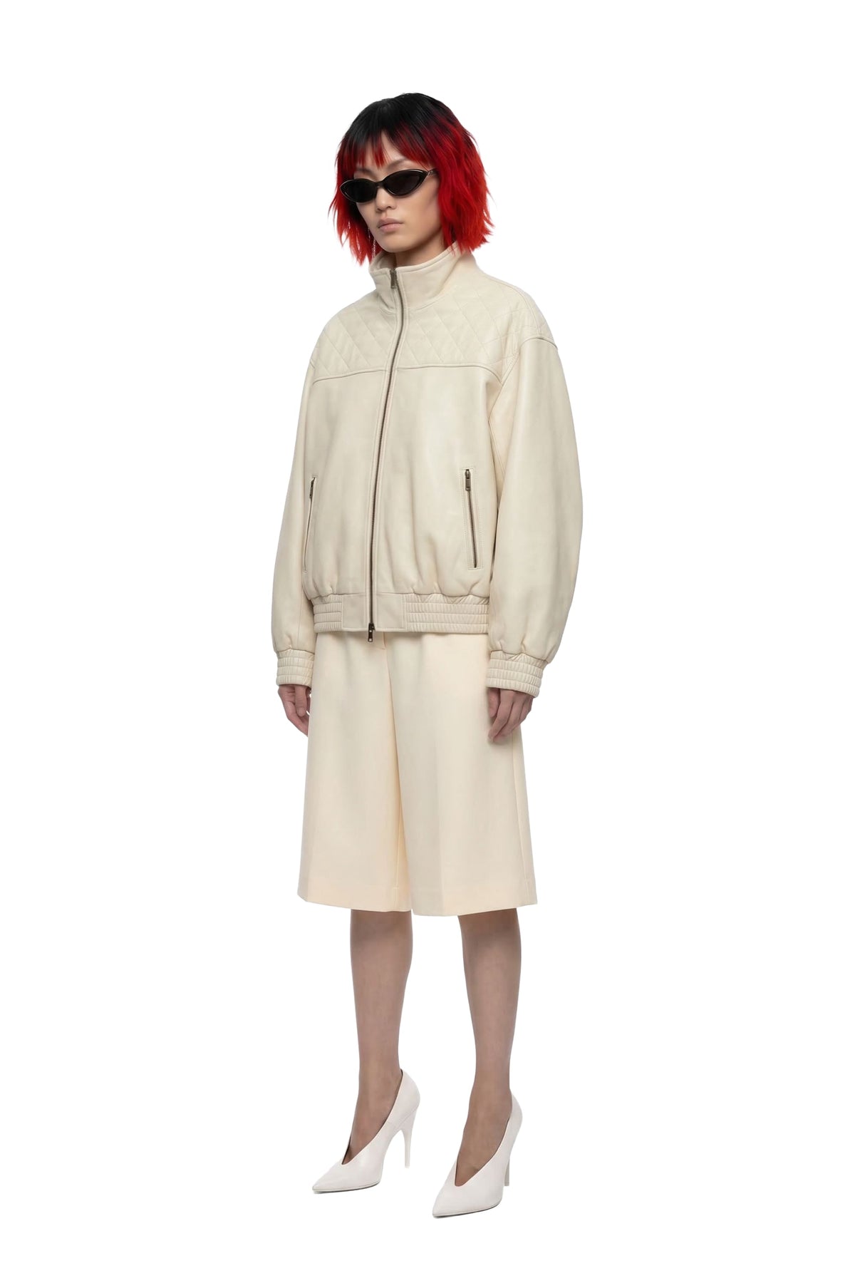 A model with short red hair and black sunglasses wears a cream-colored two-piece jacket and skirt set with white pointed-toe heels, set against a plain white background.
