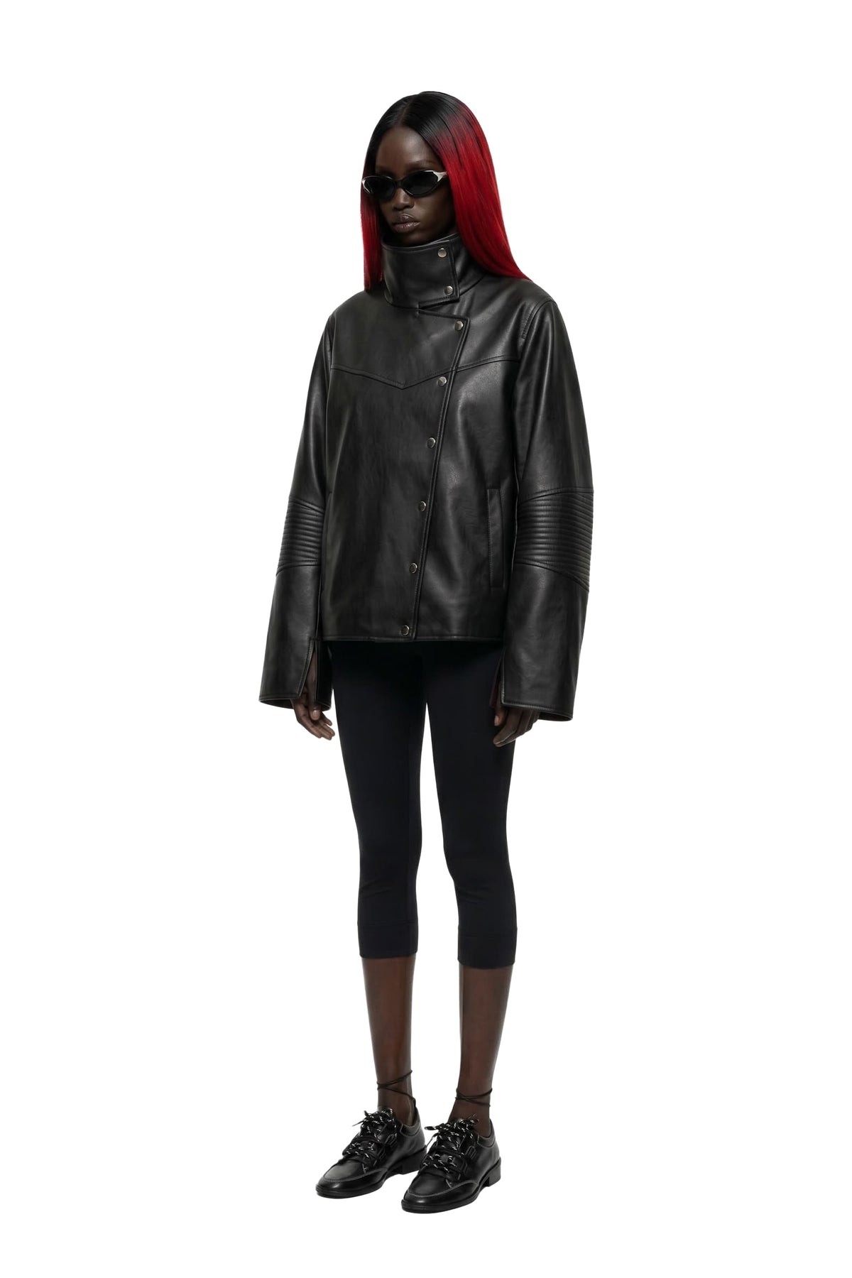 A model with long red hair and black sunglasses wearing a black leather jacket, black cropped leggings, and black lace-up shoes, standing against a solid white background.