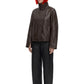 A model with a bright orange-red bob haircut and black sunglasses stands against a white background, wearing a dark brown leather jacket with a high mock neck, black wide-leg trousers, and black shoes.