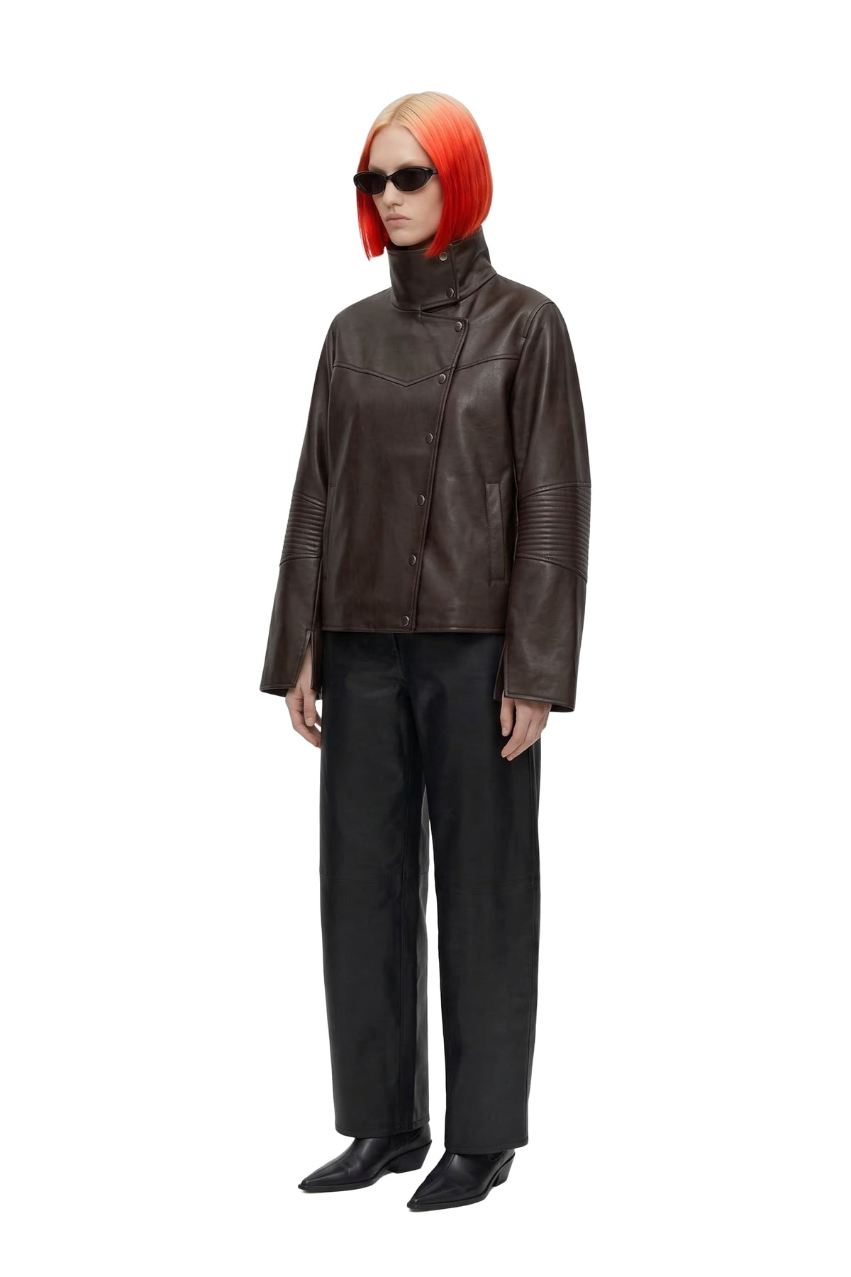 A model with a bright orange-red bob haircut and black sunglasses stands against a white background, wearing a dark brown leather jacket with a high mock neck, black wide-leg trousers, and black shoes.
