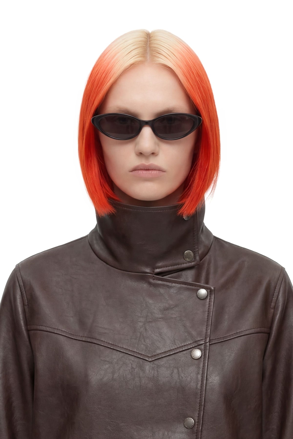 A model with a short, bright orange-red ombre blonde bob hairstyle, wearing black cat-eye sunglasses and a dark brown leather jacket, set against a plain white background.