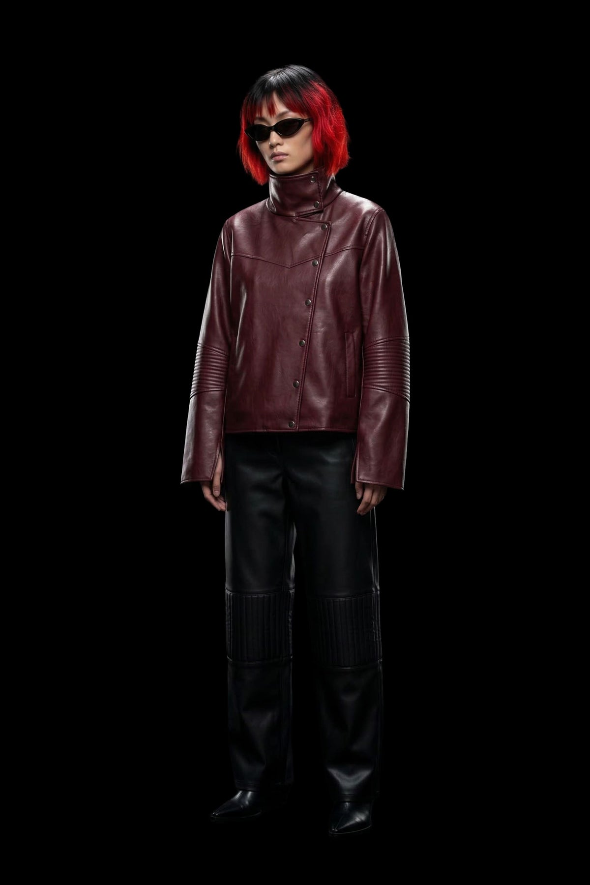 A model with a short red and black bob haircut, wearing black sunglasses, a dark burgundy leather jacket, and black leather pants, standing against a solid black background.