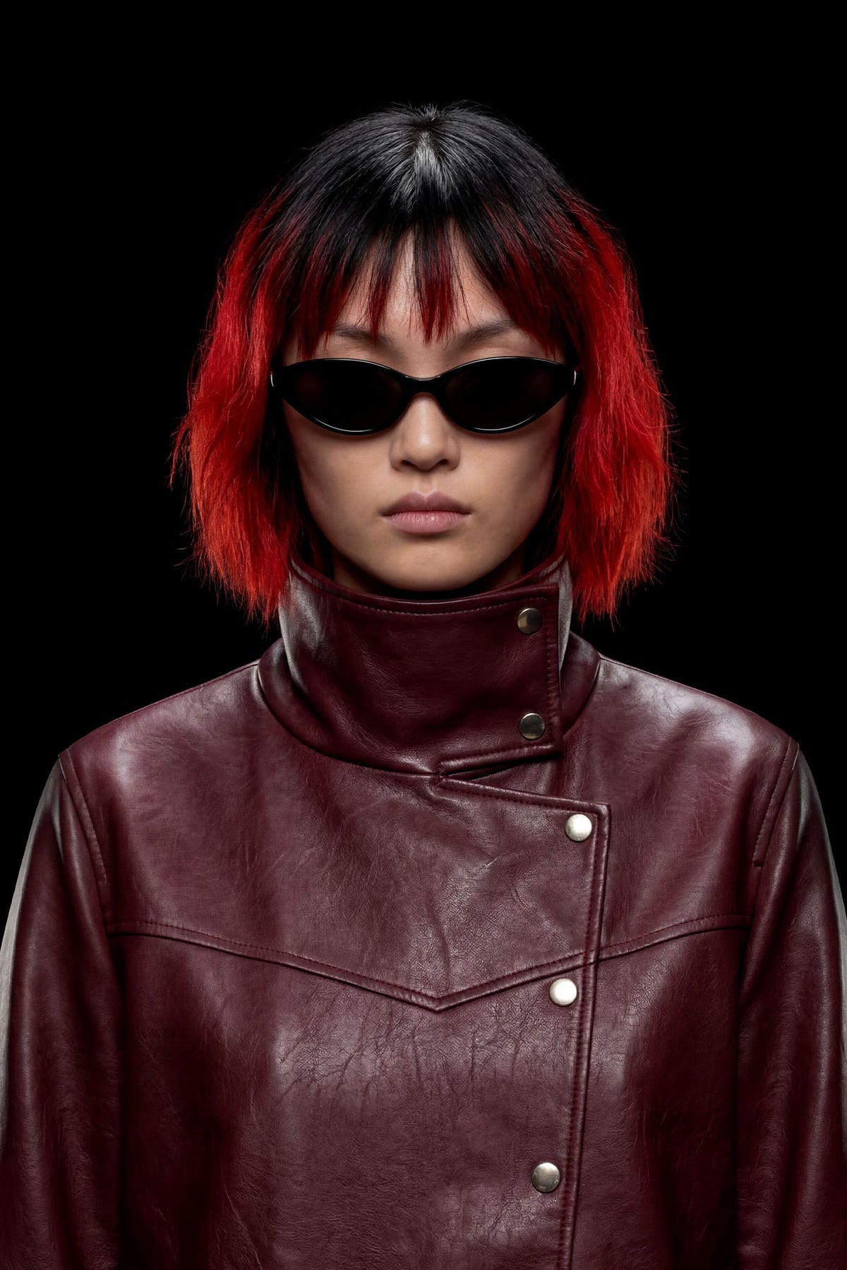 A model with a short black and vibrant red ombre wig, wearing black cat-eye sunglasses and a high-collared burgundy leather jacket, set against a solid black background.