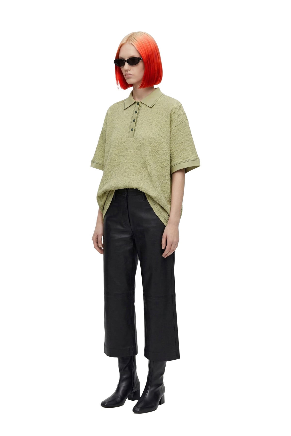 A model with a bright orange-red bob haircut and black sunglasses is shown from the head to the boots, wearing an oversized light olive green short-sleeve polo shirt, black wide-leg cropped trousers, and black ankle boots, against a plain white background.