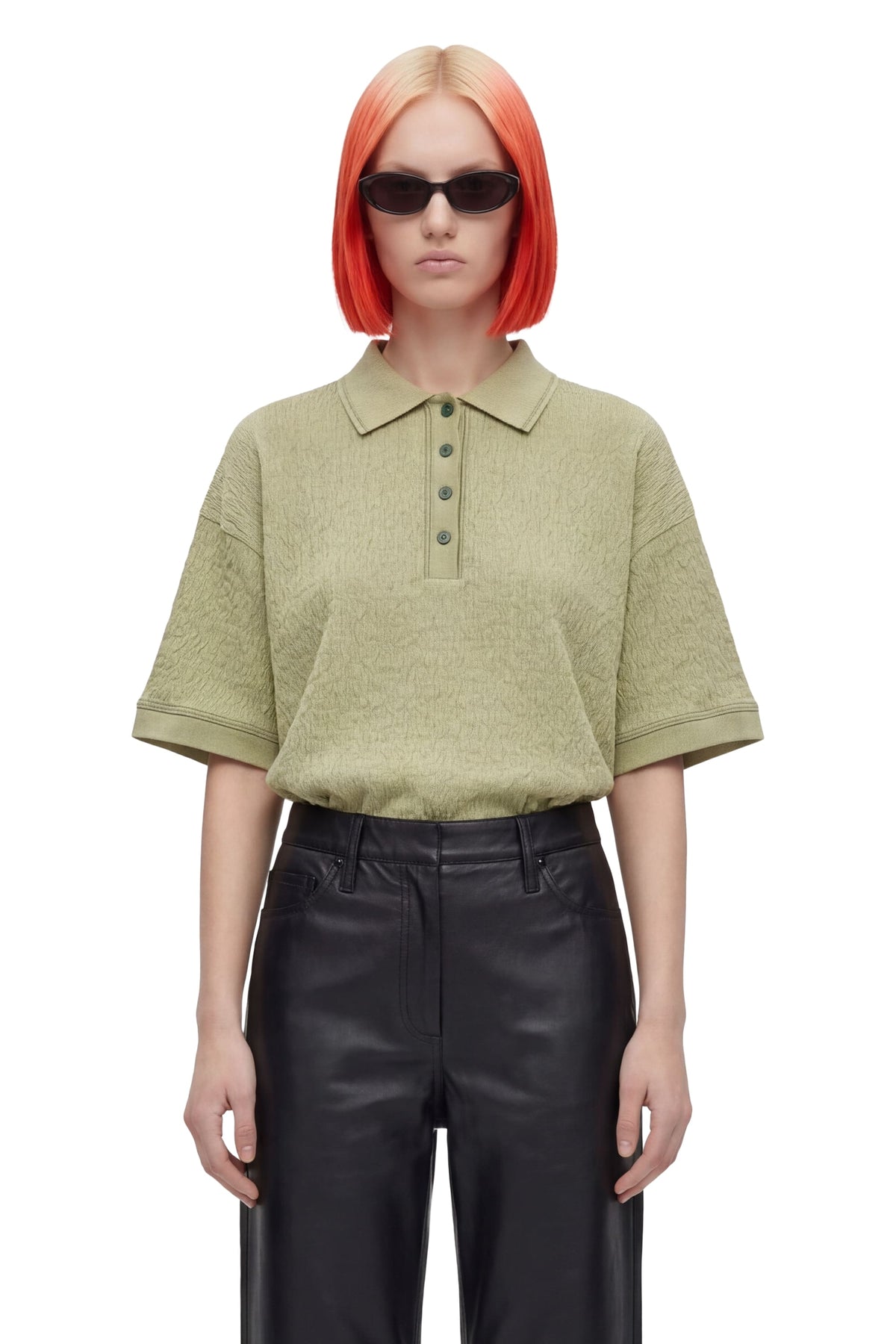 A model with bright orange hair and sunglasses wearing a short-sleeve olive green knit polo shirt and black leather pants, shot against a solid white background.