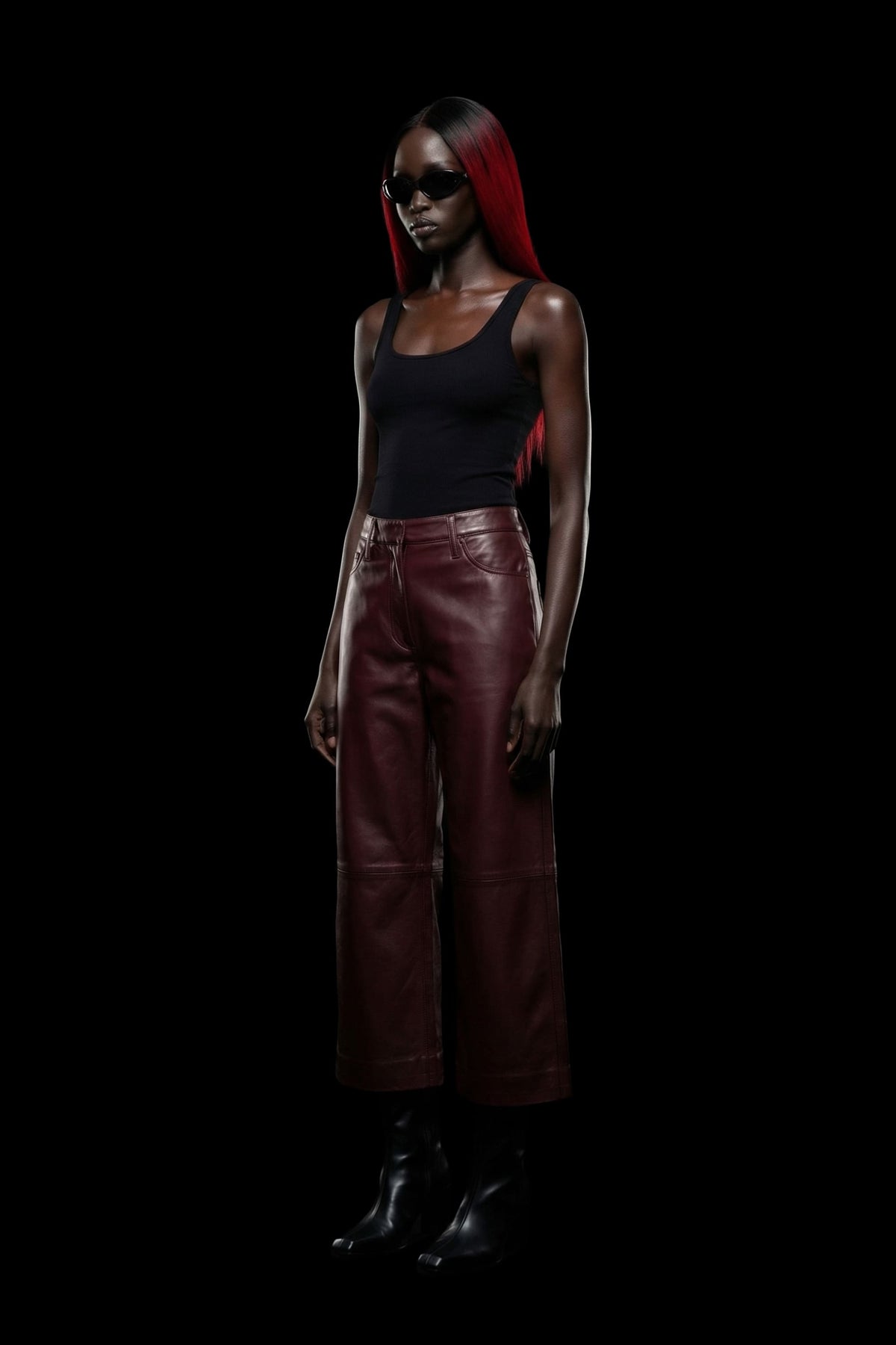 A model with long red hair stands against a solid black background, wearing a black tank top, high-waisted dark red leather wide-leg pants, and black boots.