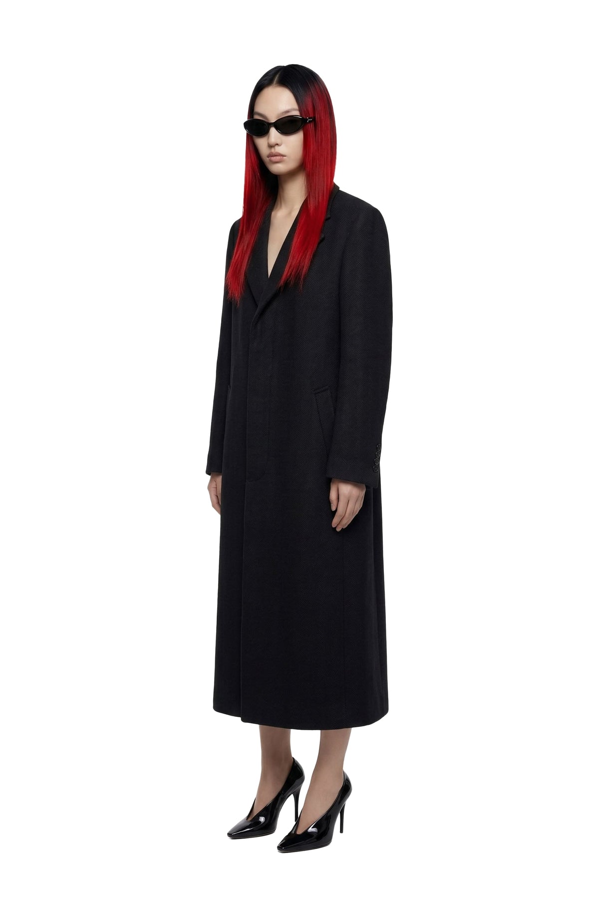 A model with long red hair wearing a long black coat, black sunglasses, and black patent leather high heels, standing against a solid white background.