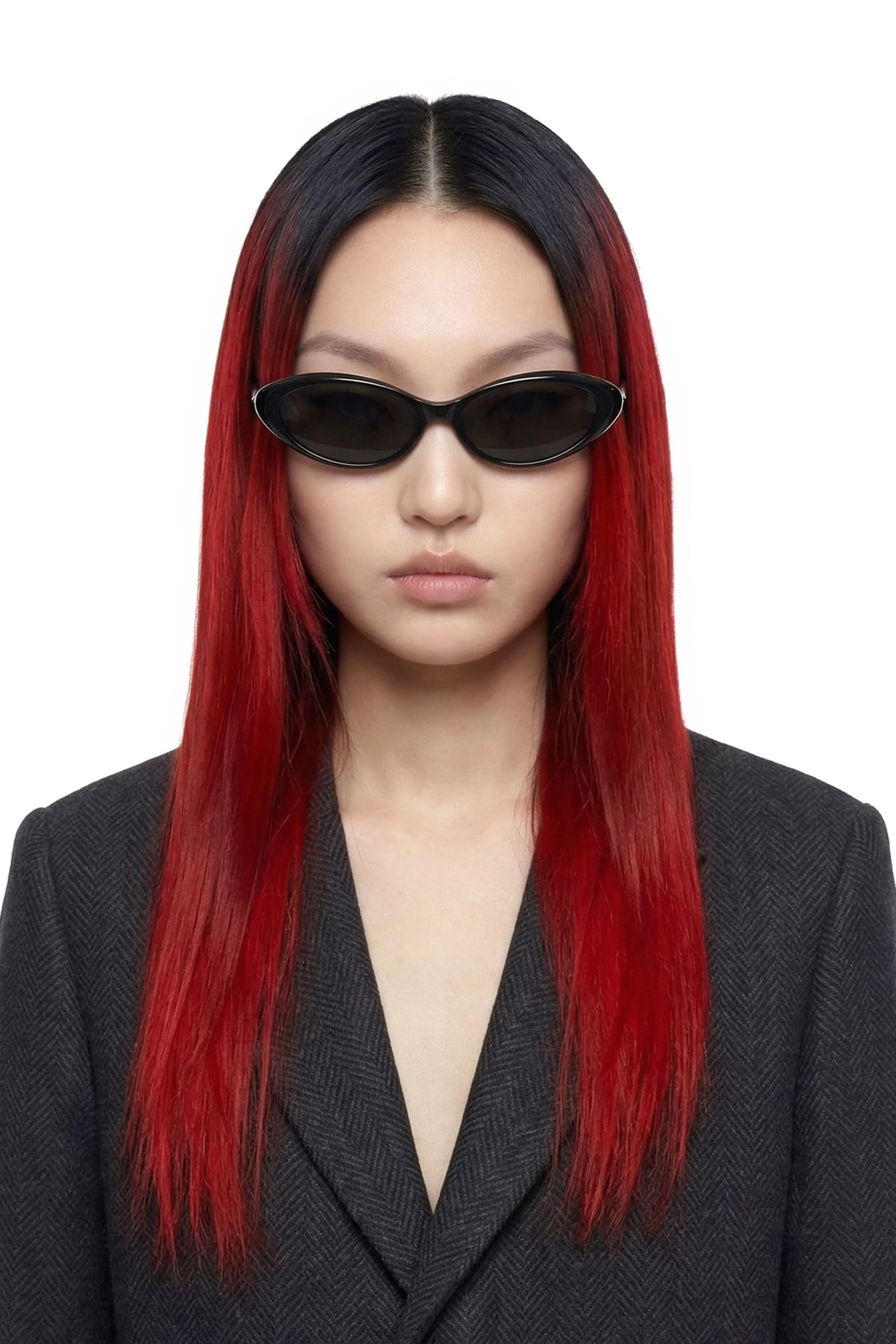 A model with long, straight black hair featuring vibrant red ombre ends, wearing black oval sunglasses and a dark gray blazer against a plain white background.