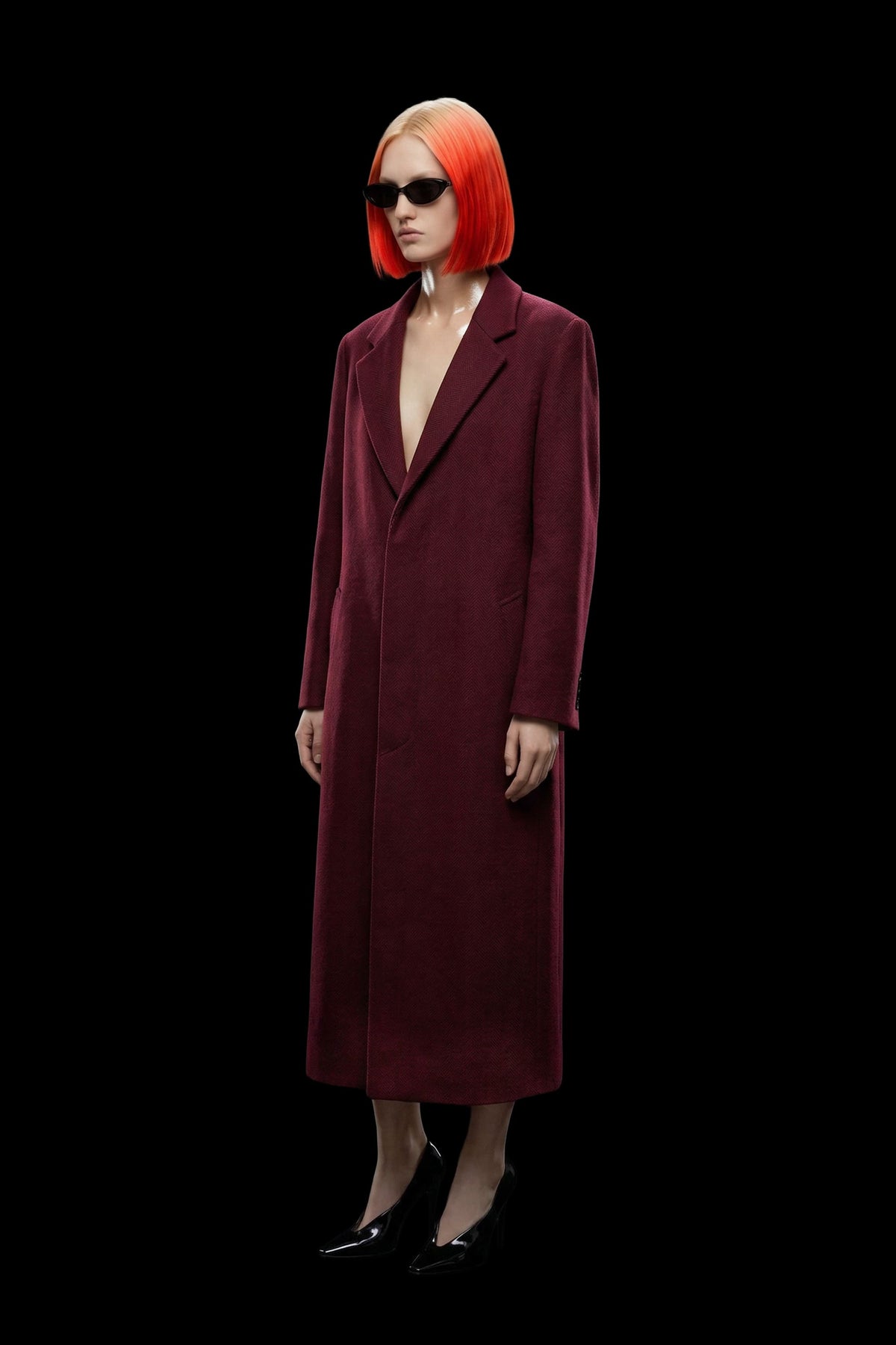 A model with a vibrant orange-red bob haircut and black sunglasses wears a long, deep burgundy wool-blend coat with black pointed-toe heels, set against a solid black background.