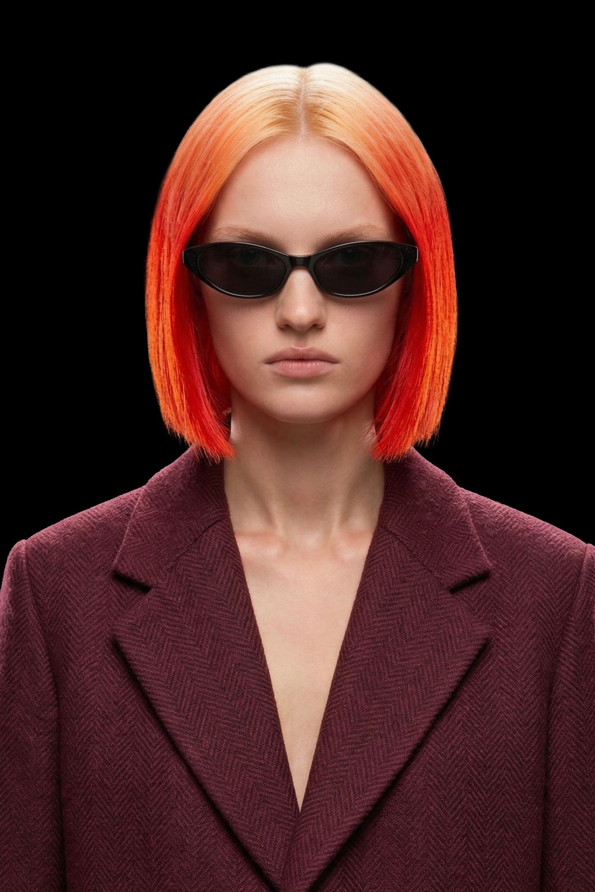 A model with a vibrant orange-to-red ombre bob haircut wearing black sunglasses and a textured burgundy blazer, set against a solid black background.
