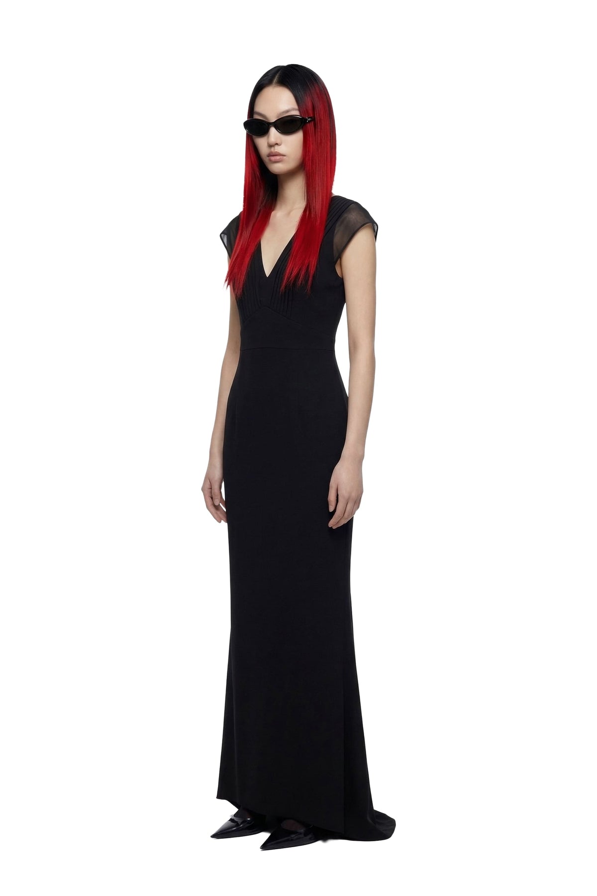 A model with long red hair and black sunglasses wearing a floor-length black dress with sheer cap sleeves, photographed against a plain white background.