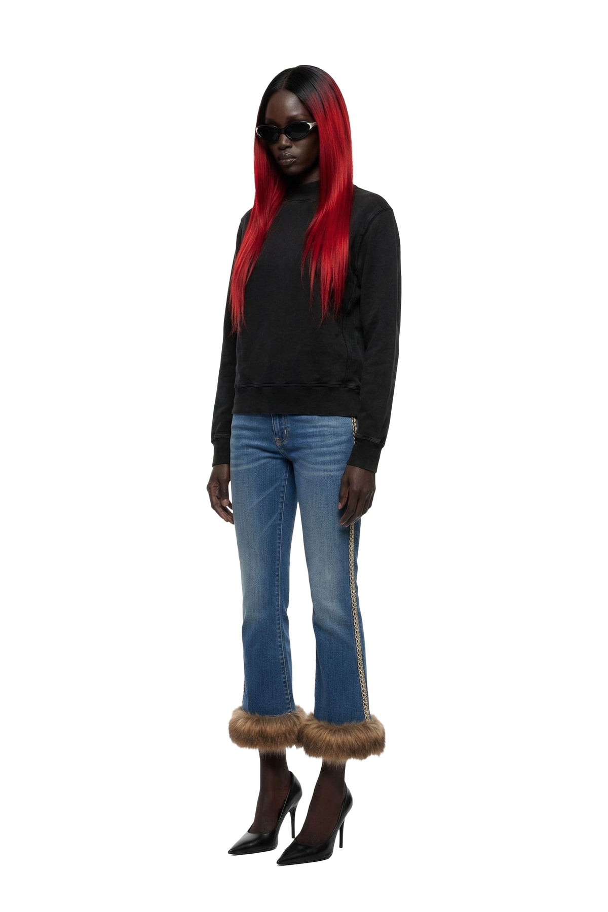A full-body model with long red-and-black hair wearing a black long-sleeve top, blue jeans with brown faux-fur trim at the hem, and black pointed-toe heels against a plain white background.