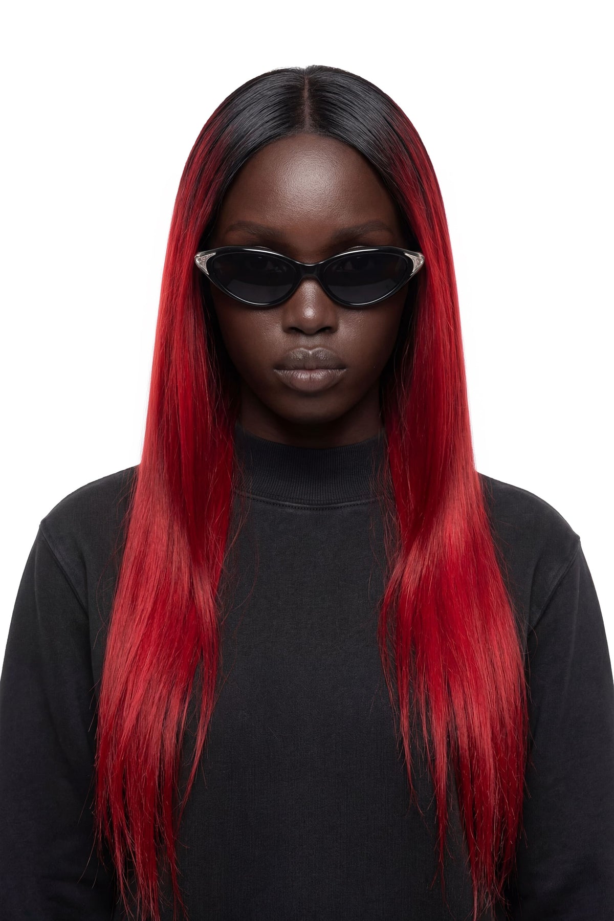 A model with long, straight black hair featuring vibrant red ombre ends, wearing black cat-eye sunglasses and a black turtleneck against a solid white background.