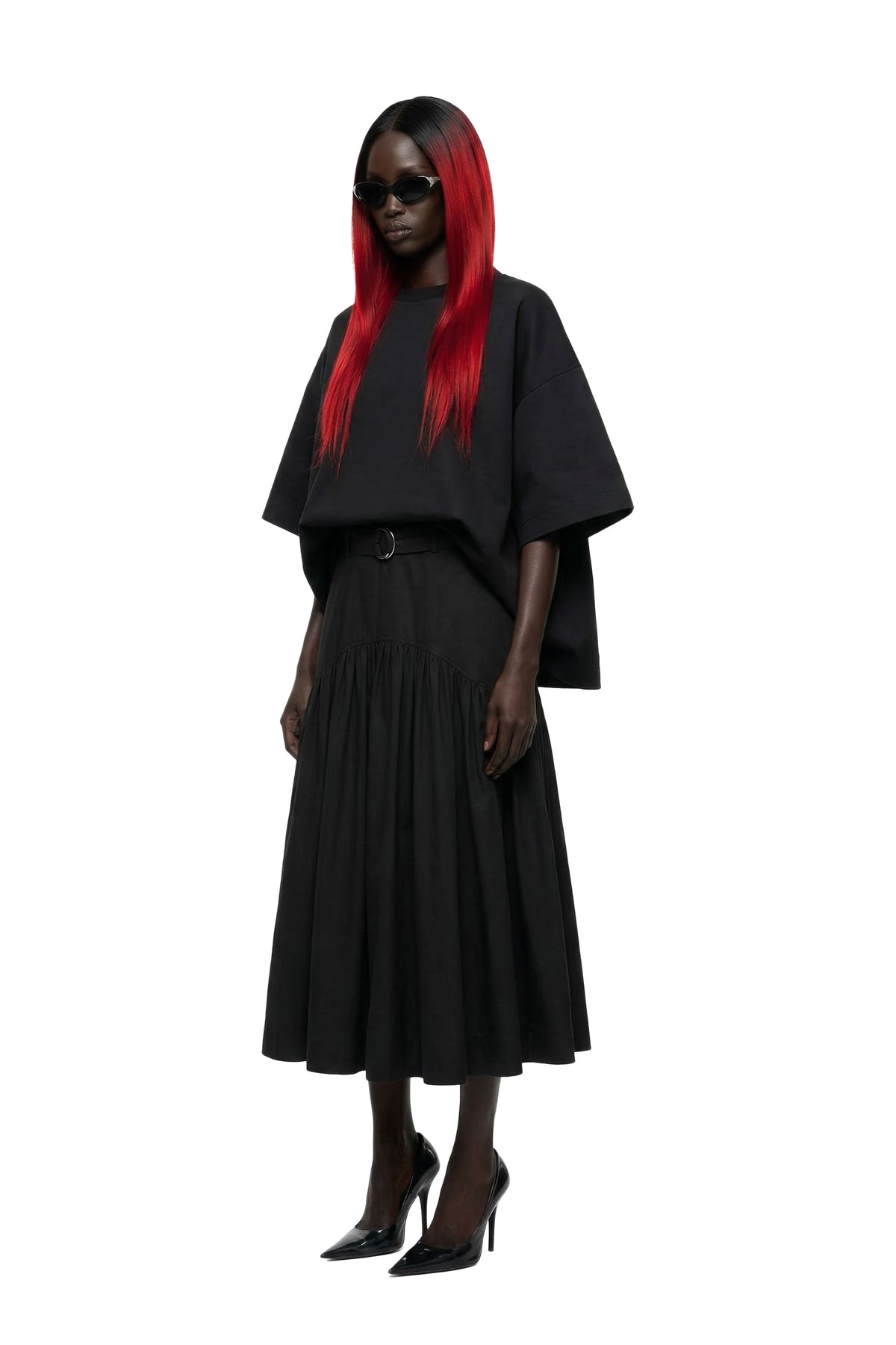 A model with long red hair wearing a black draped short-sleeve top, a black pleated midi skirt, and black pointed-toe heels against a solid white background.