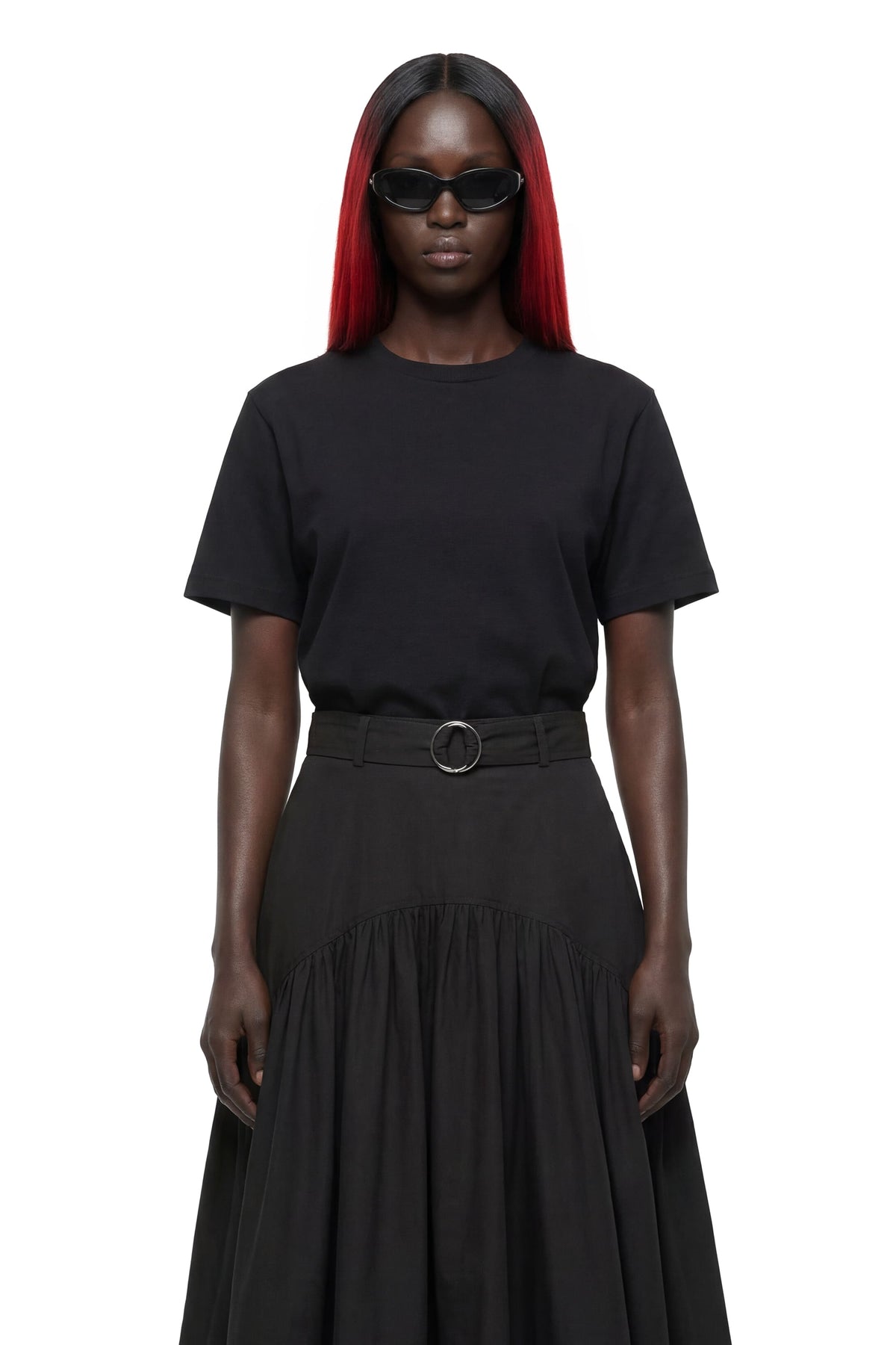 A model with long red hair wearing a black short-sleeve crewneck top and a matching black pleated skirt with a belt, photographed against a solid white background.