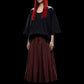 A model with long, straight red hair and black sunglasses is shown from the waist down, wearing a loose black top and a pleated dark red skirt, set against a solid black background.